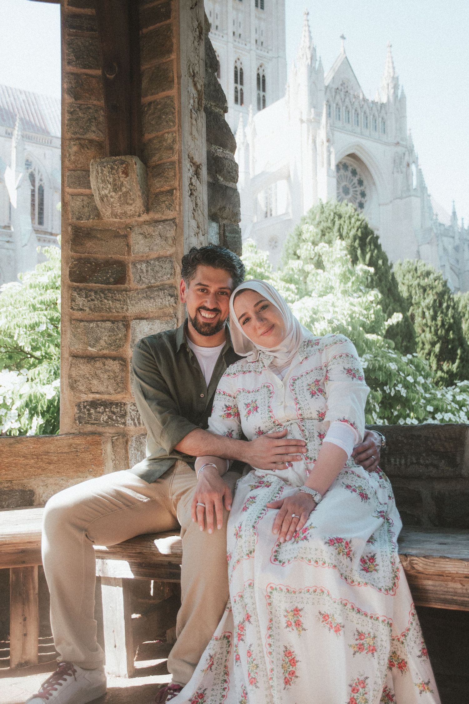 A smiling couple sits together outdoors near a stone column with a Gothic cathedral in the background.