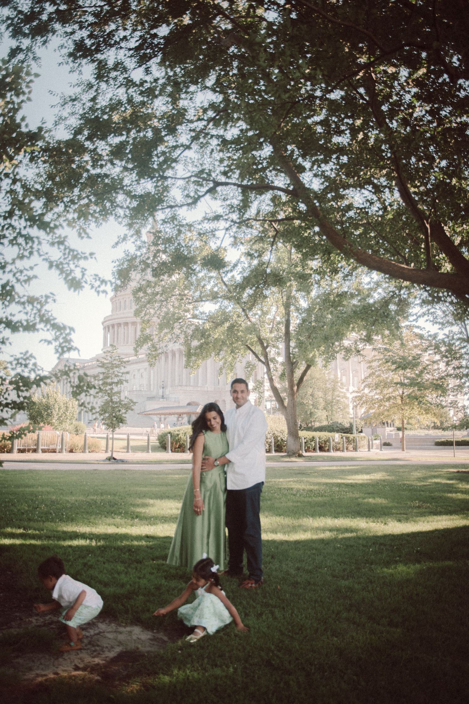 Family portrait on green lawn with large tree and government building backdrop, parents in green and white attire.