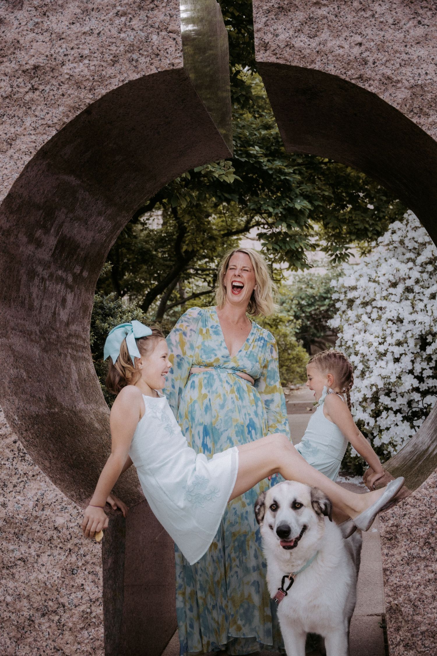 A laughing woman in a floral dress with two children and a white dog inside a circular stone sculpture.