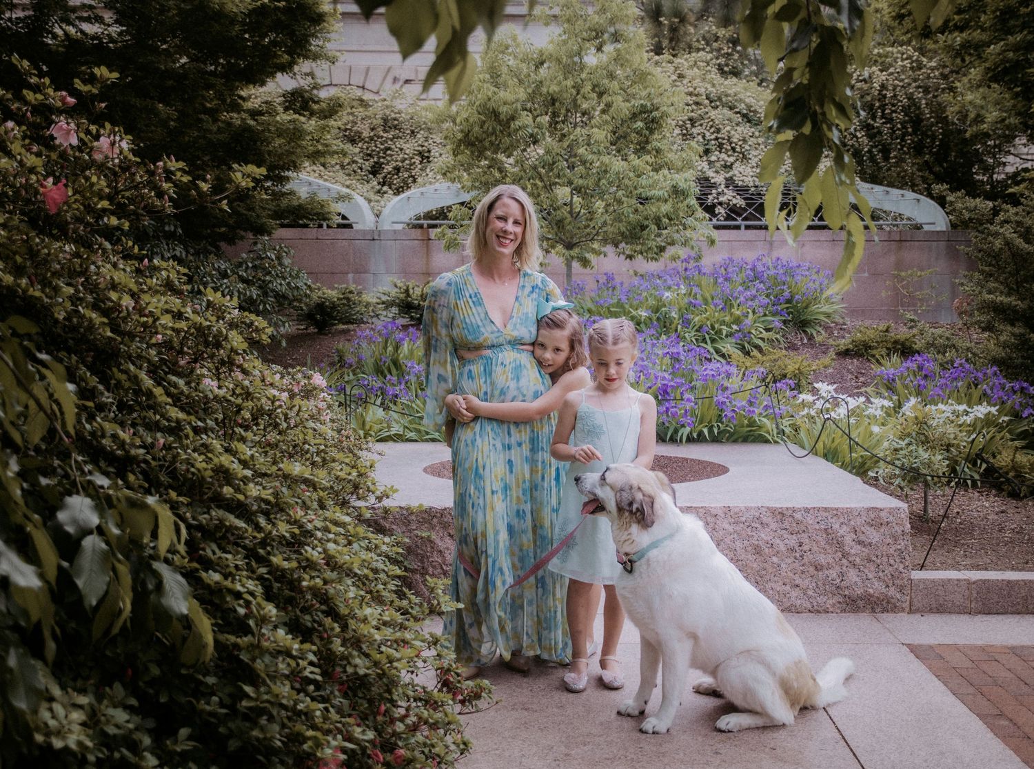 Woman in floral dress poses with two girls and a large white dog in a lush garden with purple flowers.