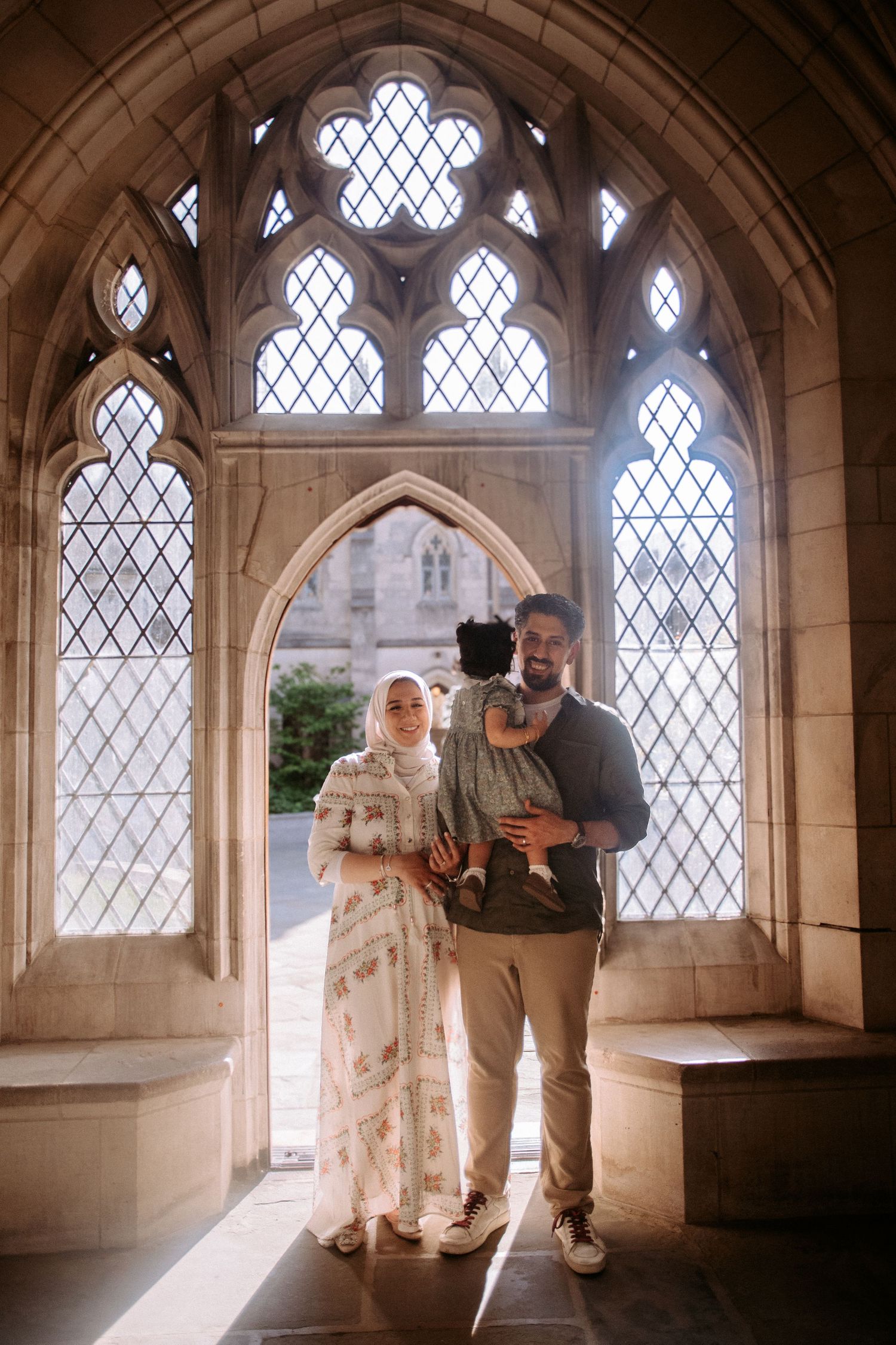Muslim family with baby posing in Gothic chapel with ornate stained glass window backdrop.