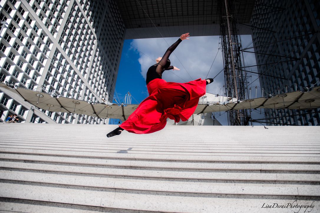 Dancer in red costume performs dramatic movements on white marble steps.
