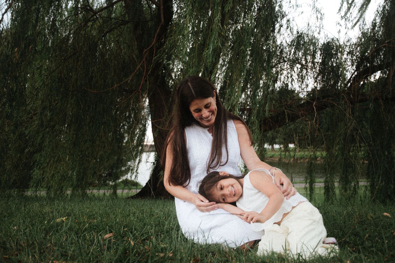 A mother and daughter share a tender moment under a weeping willow tree, sitting together on green grass near a lake.