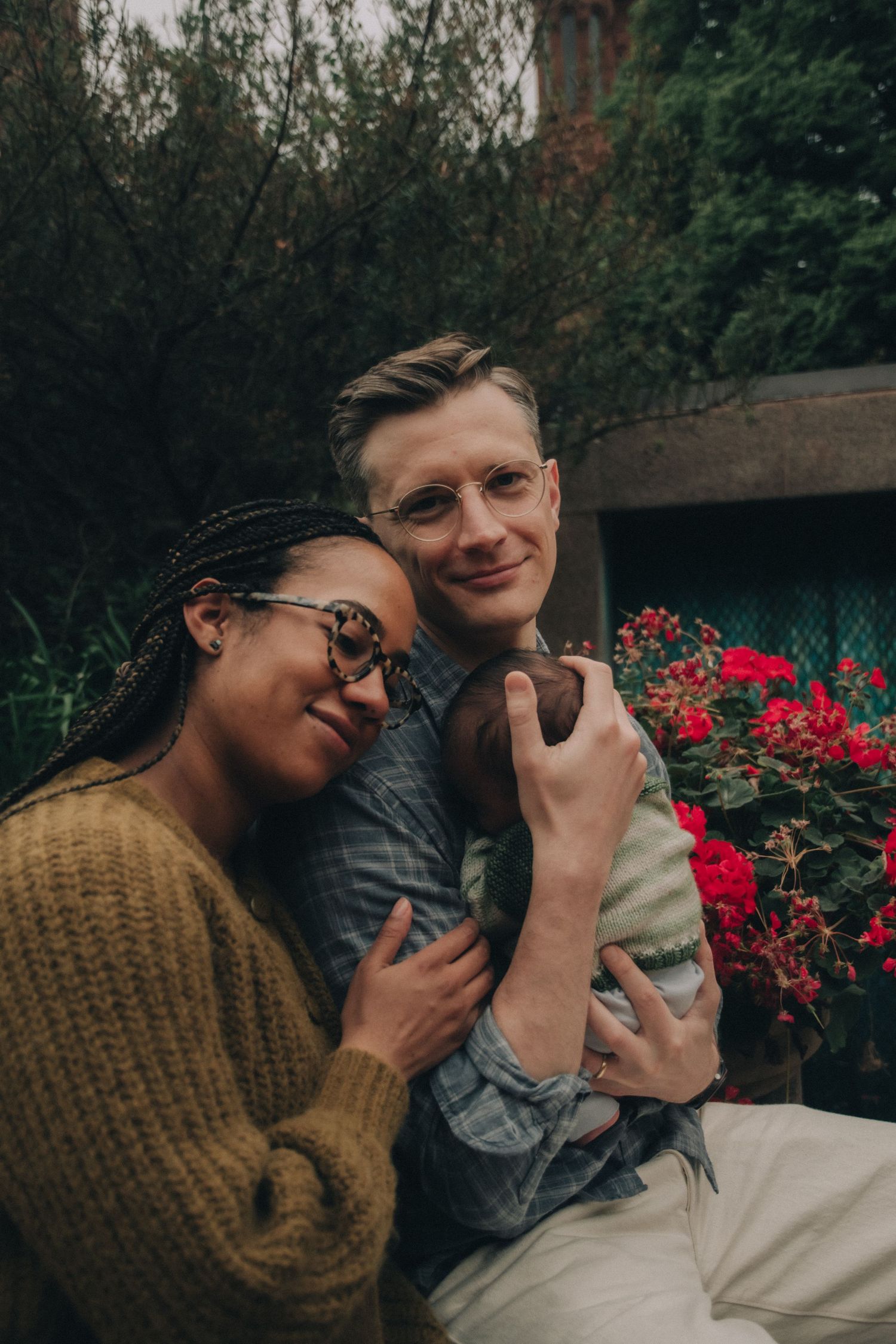 A couple wearing glasses cuddles with their newborn baby in a lush garden with red flowers.