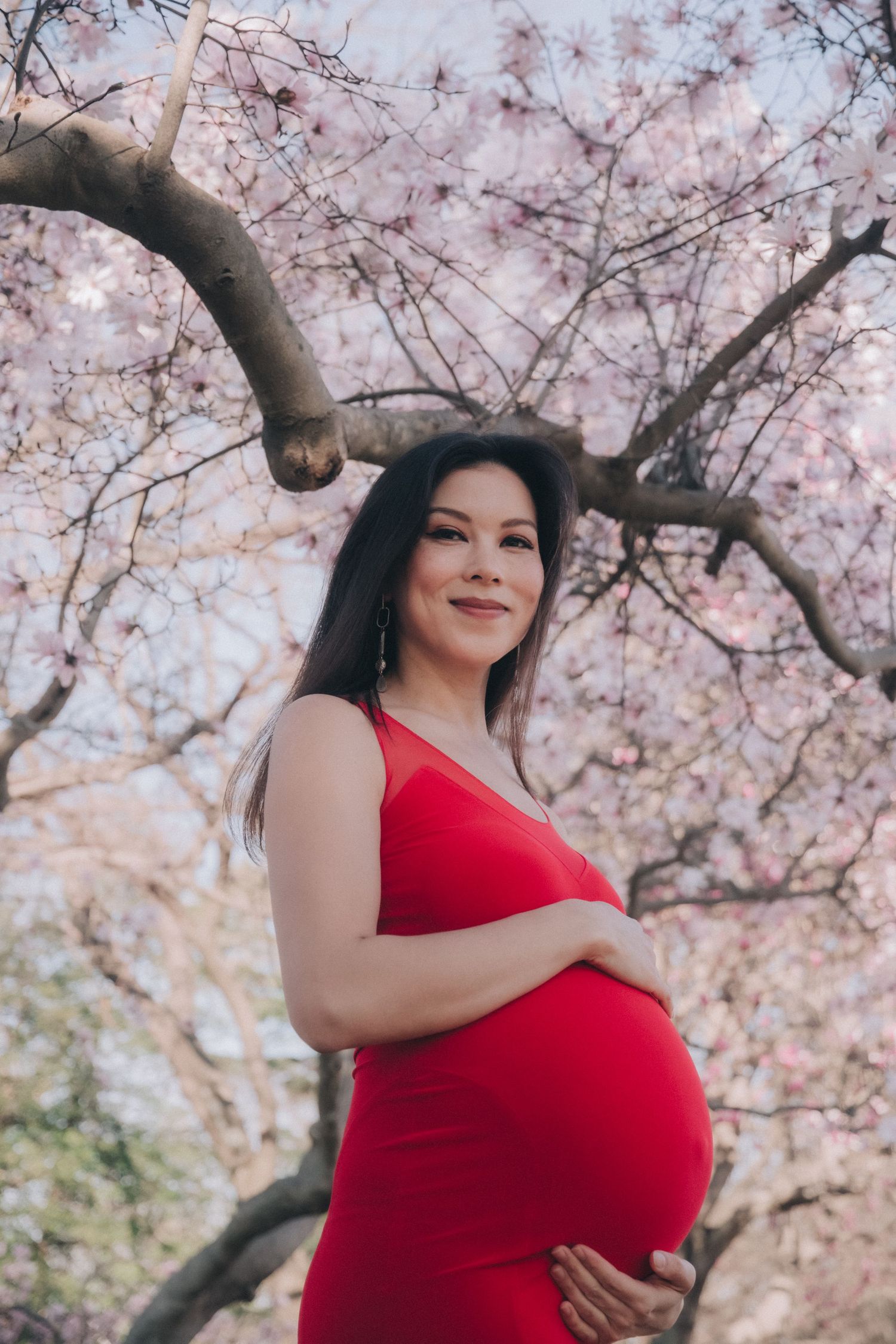 Pregnant woman in red dress cradling baby bump under pink cherry blossom tree, smiling outdoors in spring.