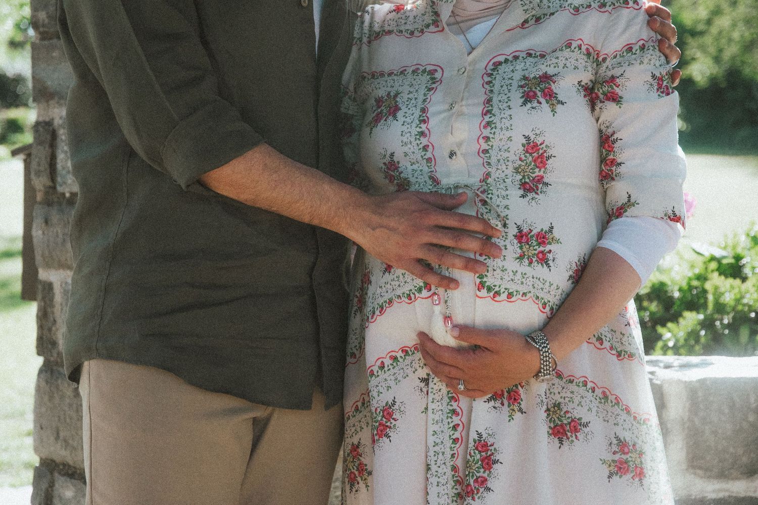 Couple tenderly cradling pregnant belly outdoors, wearing floral dress and olive shirt, maternity photo session.