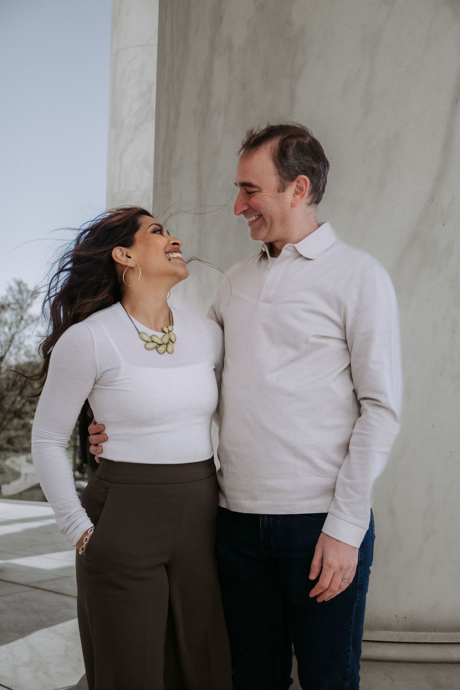 A smiling couple in white tops share a joyful moment beside marble columns outdoors.