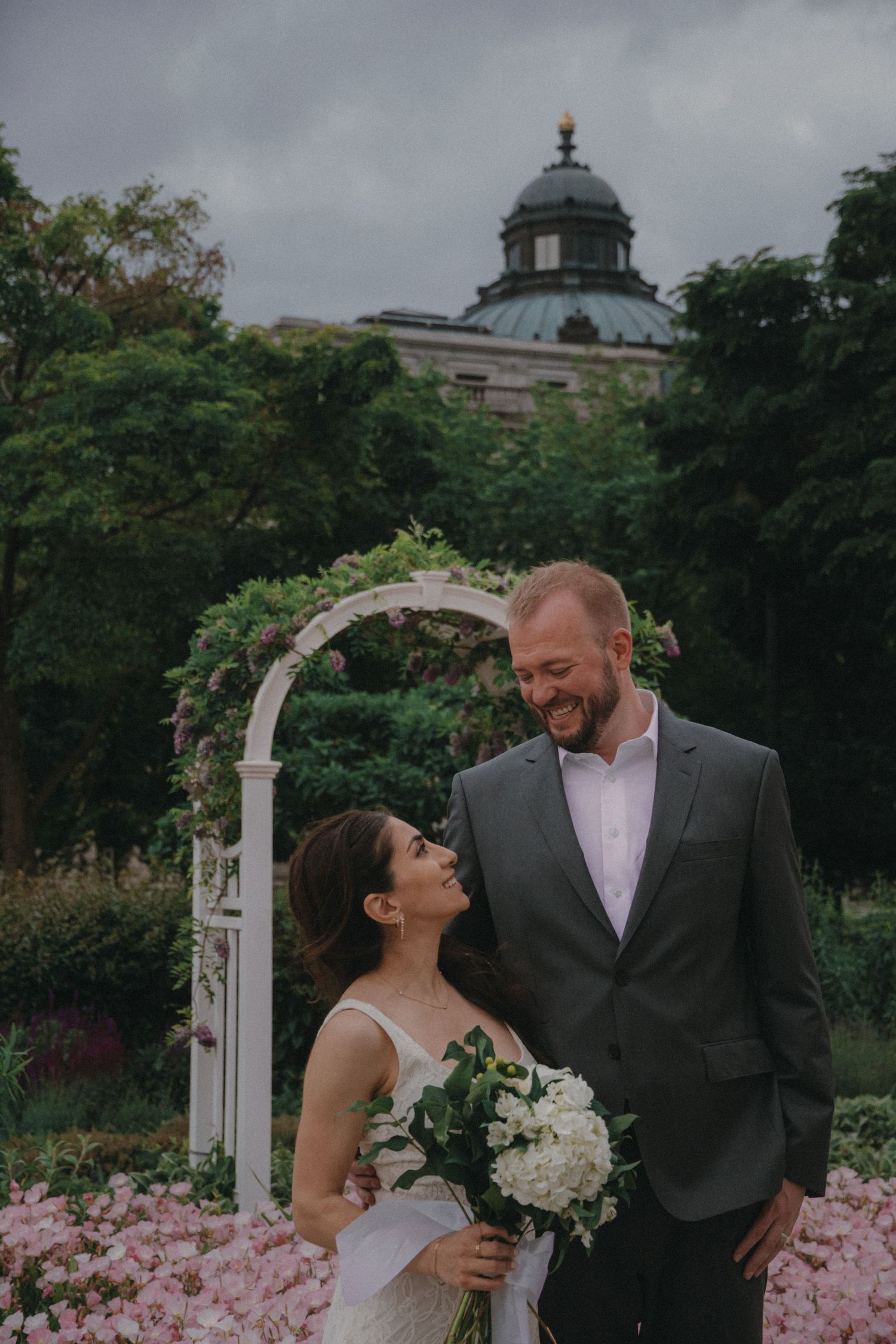 Bride and groom smile at each other in a garden with a floral arch and historic building backdrop.