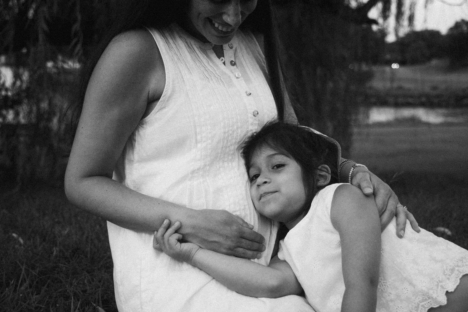 Black and white photo of a young girl hugging a pregnant woman in a white dress outdoors near trees.