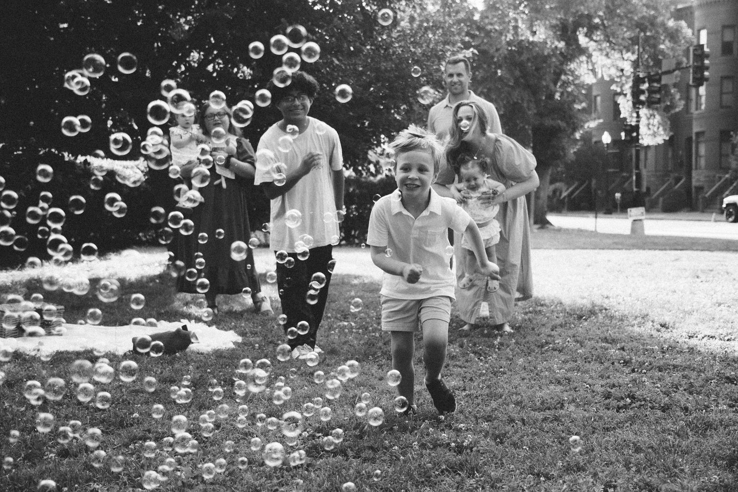 Black and white photo of children running through bubbles outdoors with family in background.