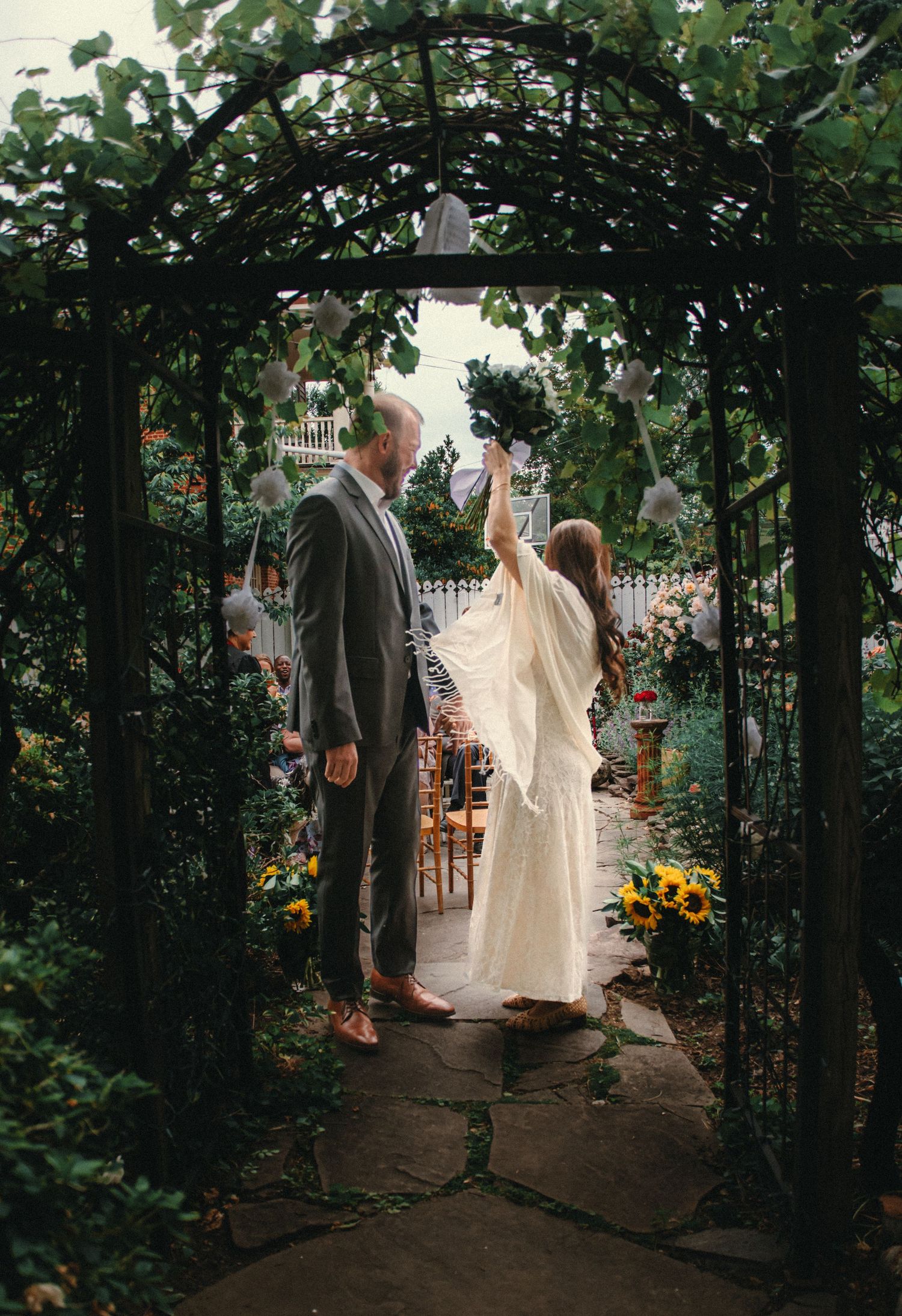Couple in wedding attire standing under a garden arch surrounded by lush greenery and sunflowers.