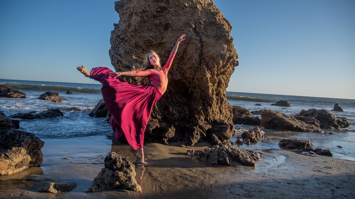 Dancer performs expressive movements along rocky beach at sunset.
