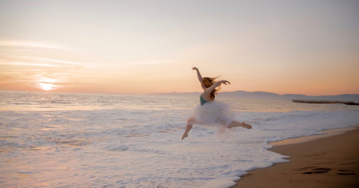 A dancer performs a graceful leap on the beach at sunset with waves crashing behind.