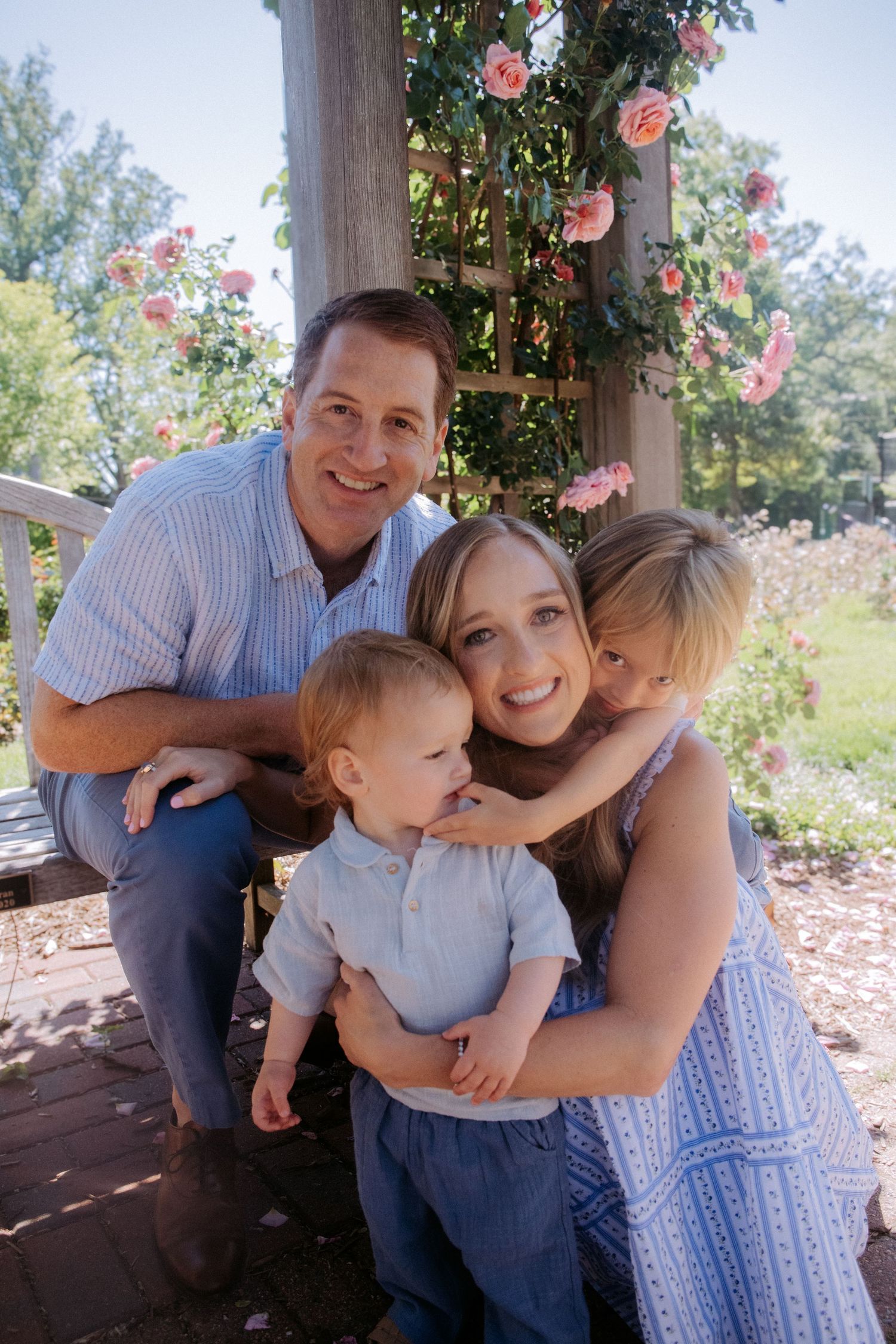 Happy family of four posing outdoors near a rose-covered pergola on a sunny day.