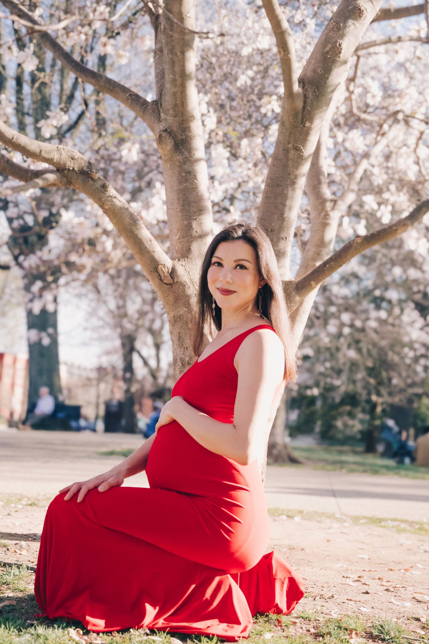 Pregnant woman in red dress sitting under blooming cherry blossom tree in park during spring.