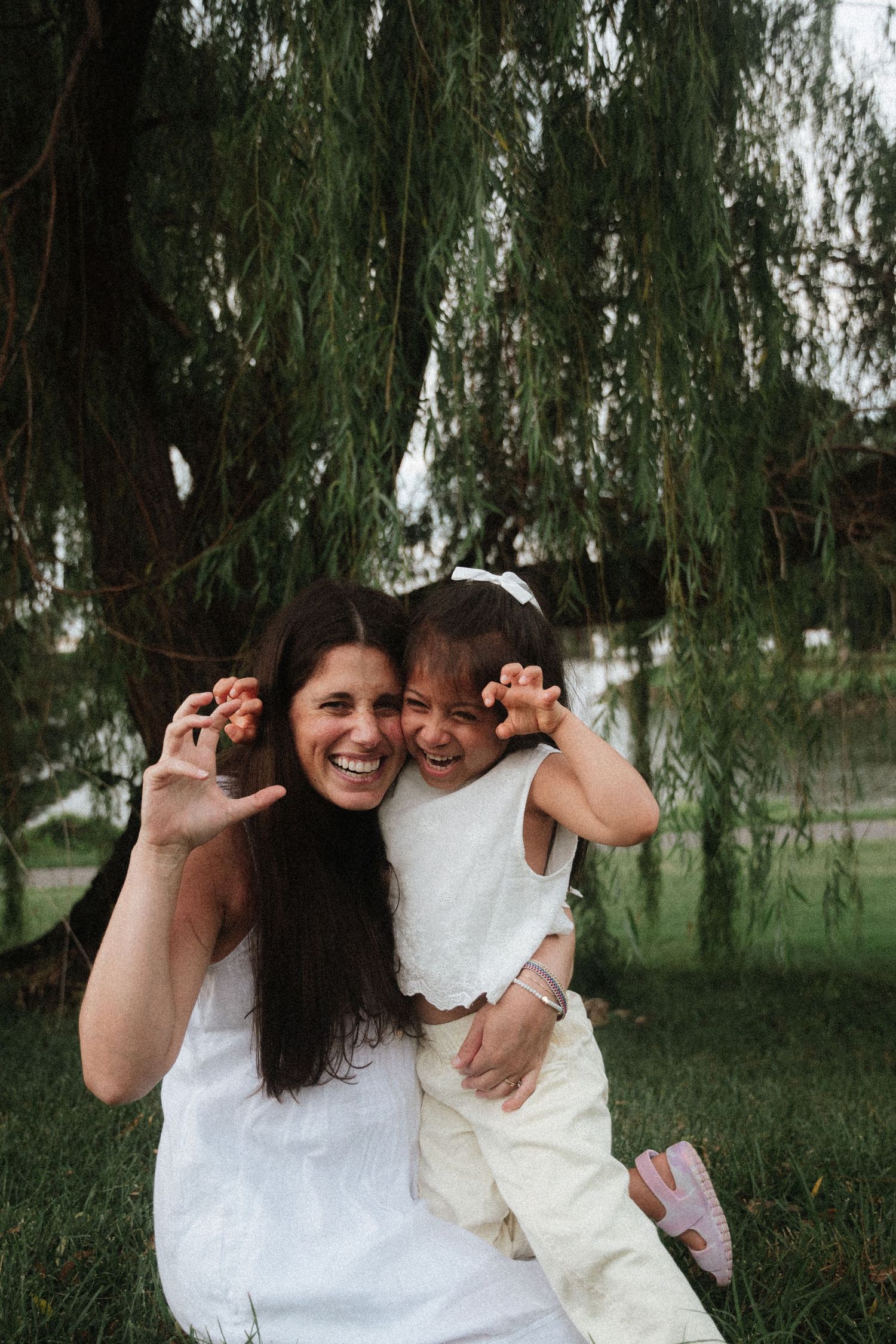 A laughing woman in white dress holds a young girl as they make silly claw gestures under a weeping willow tree.