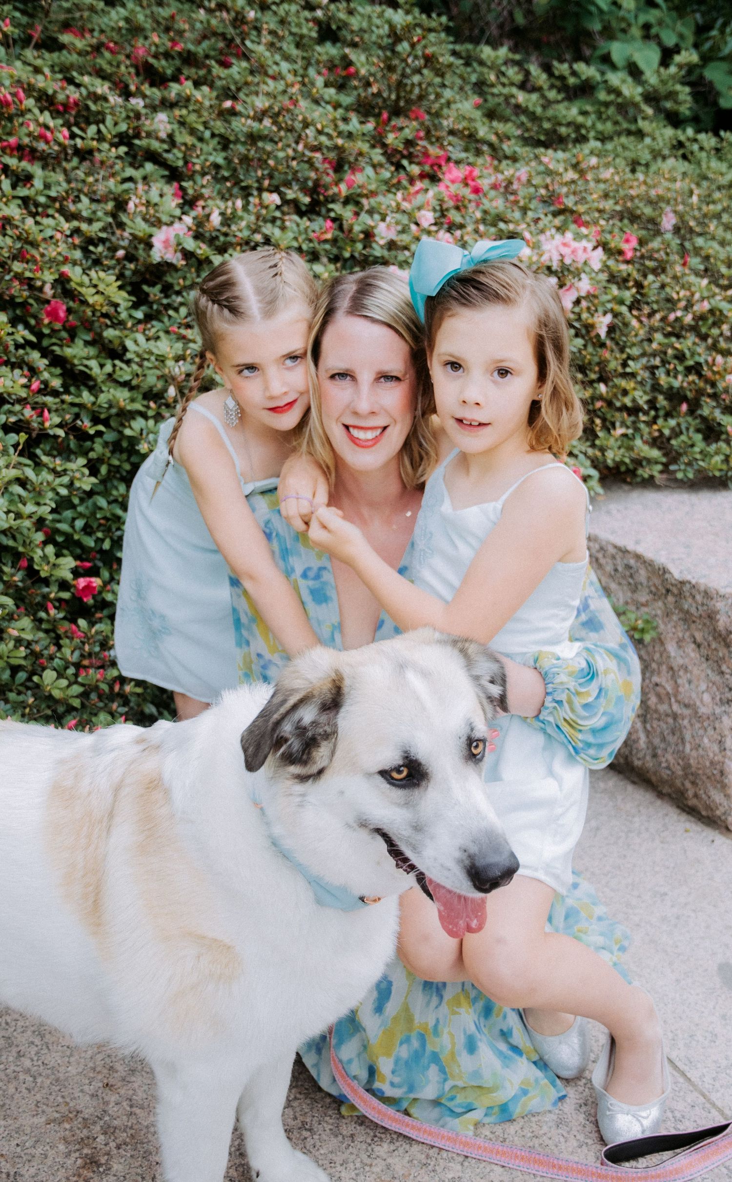 A woman poses with two young girls in pastel dresses and a large white dog in front of blooming pink flowers.