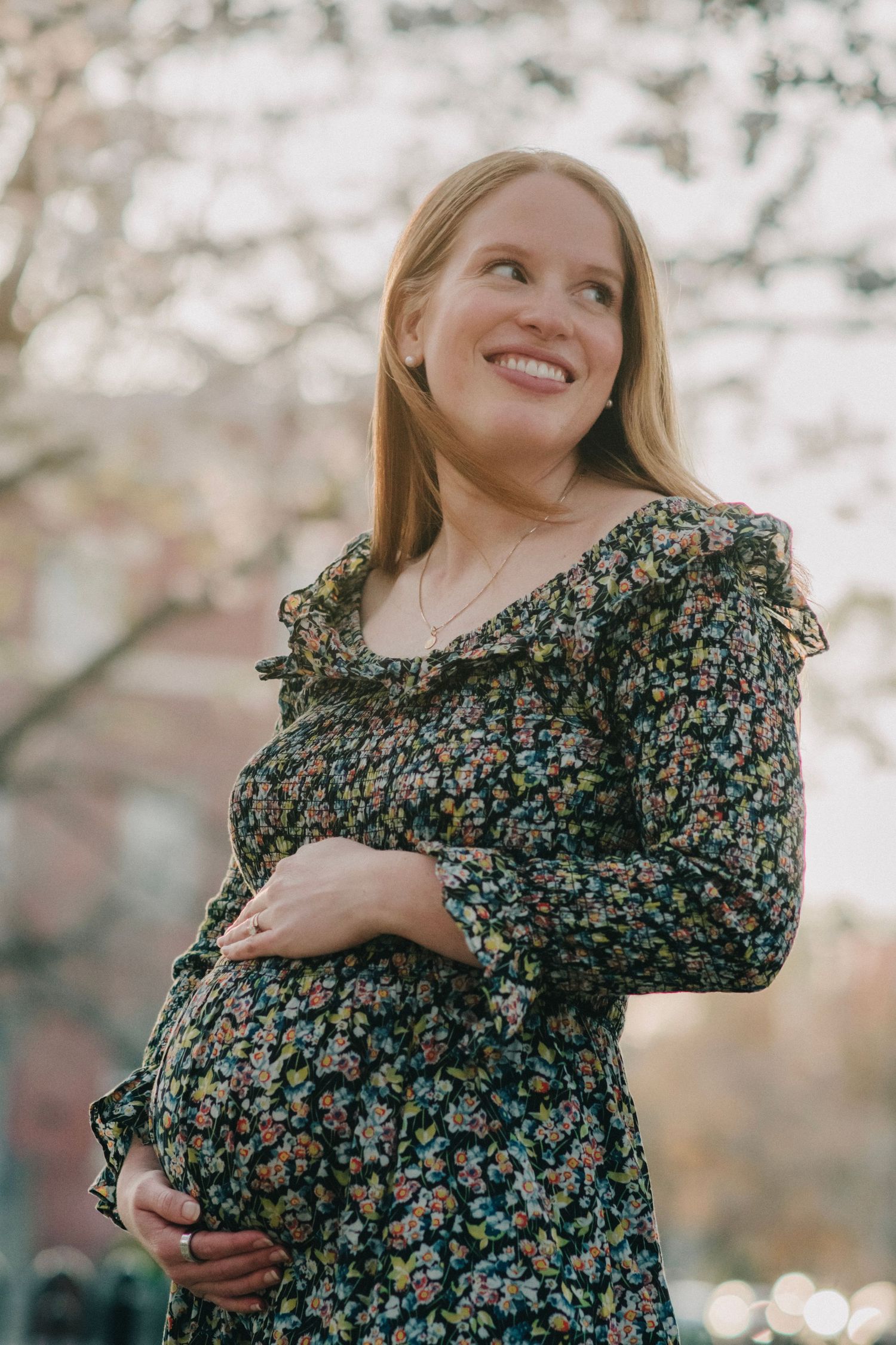 Smiling pregnant woman in floral dress cradling baby bump outdoors among spring blossoms.