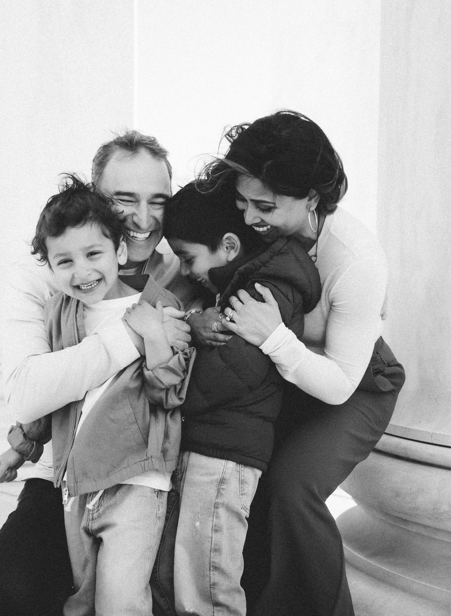 Black and white photo of a happy family of four hugging and laughing together near stone columns.