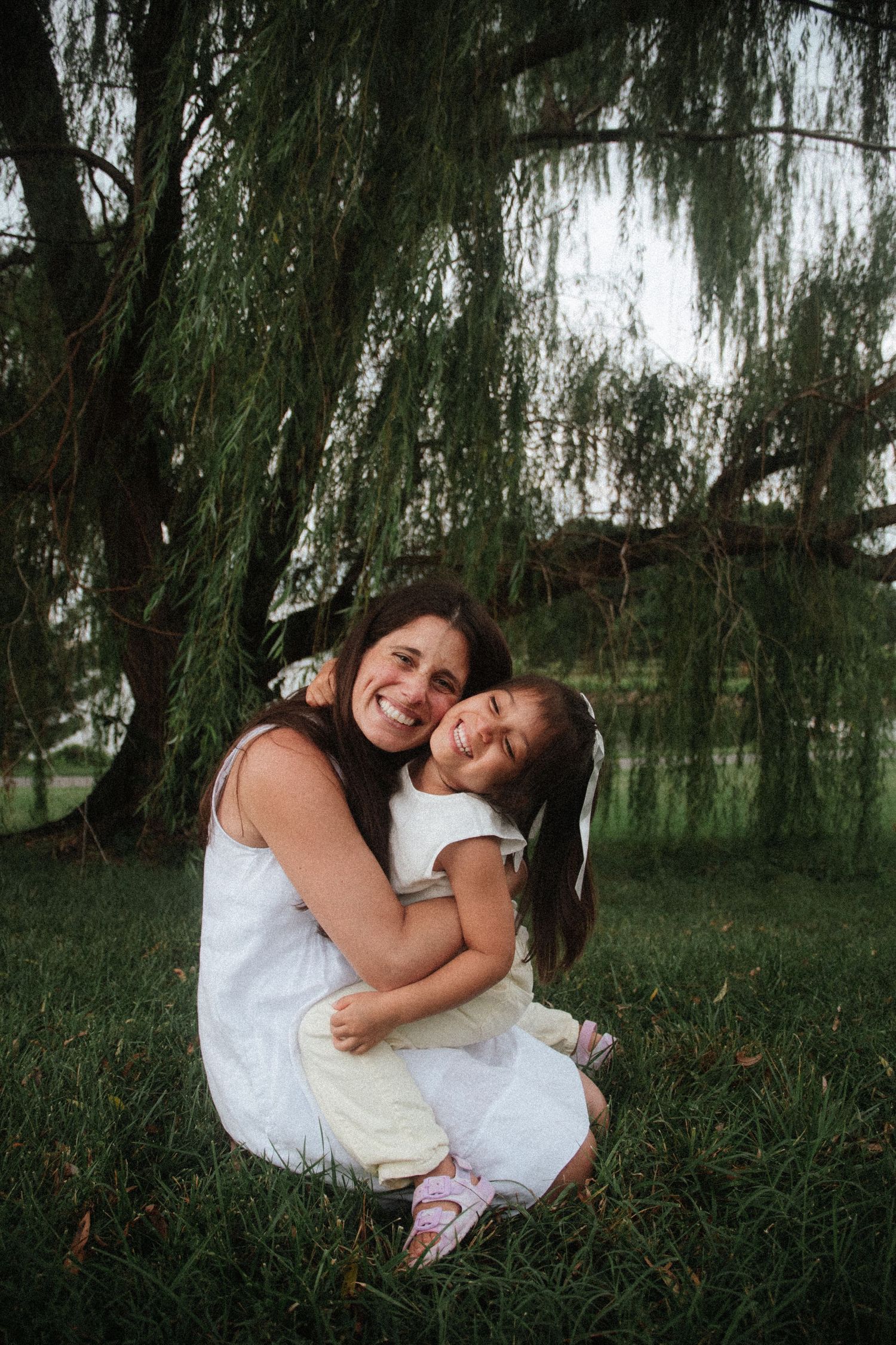 A smiling woman in white dress hugs a young girl with pigtails on green grass under a weeping willow tree.