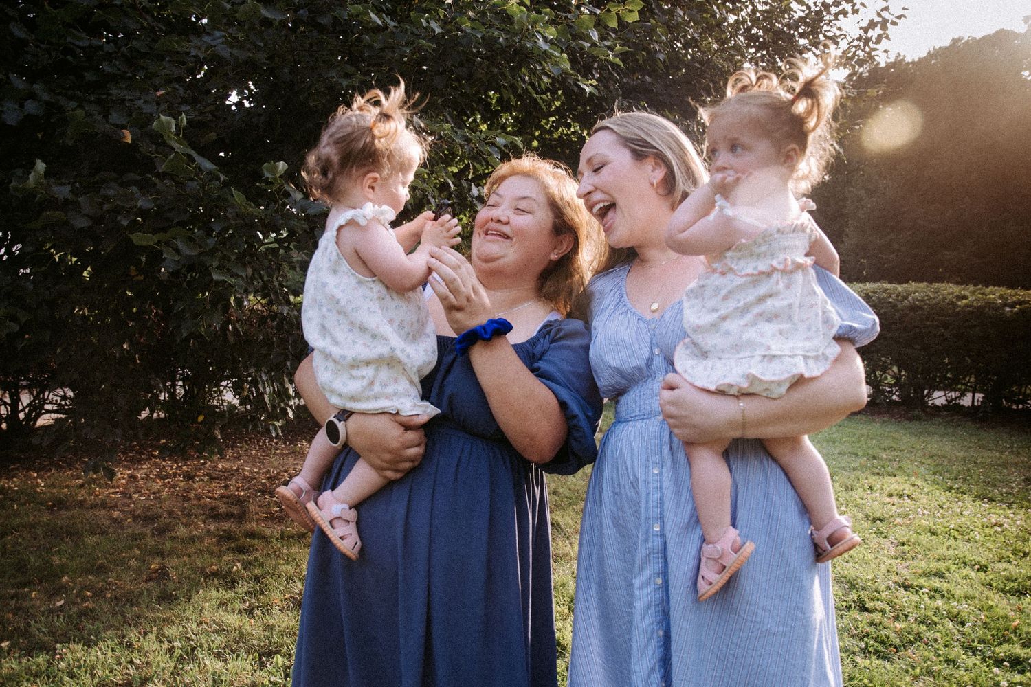Two women laughing outdoors in blue dresses, each holding a toddler girl in white floral outfits, with sunlit trees behind.
