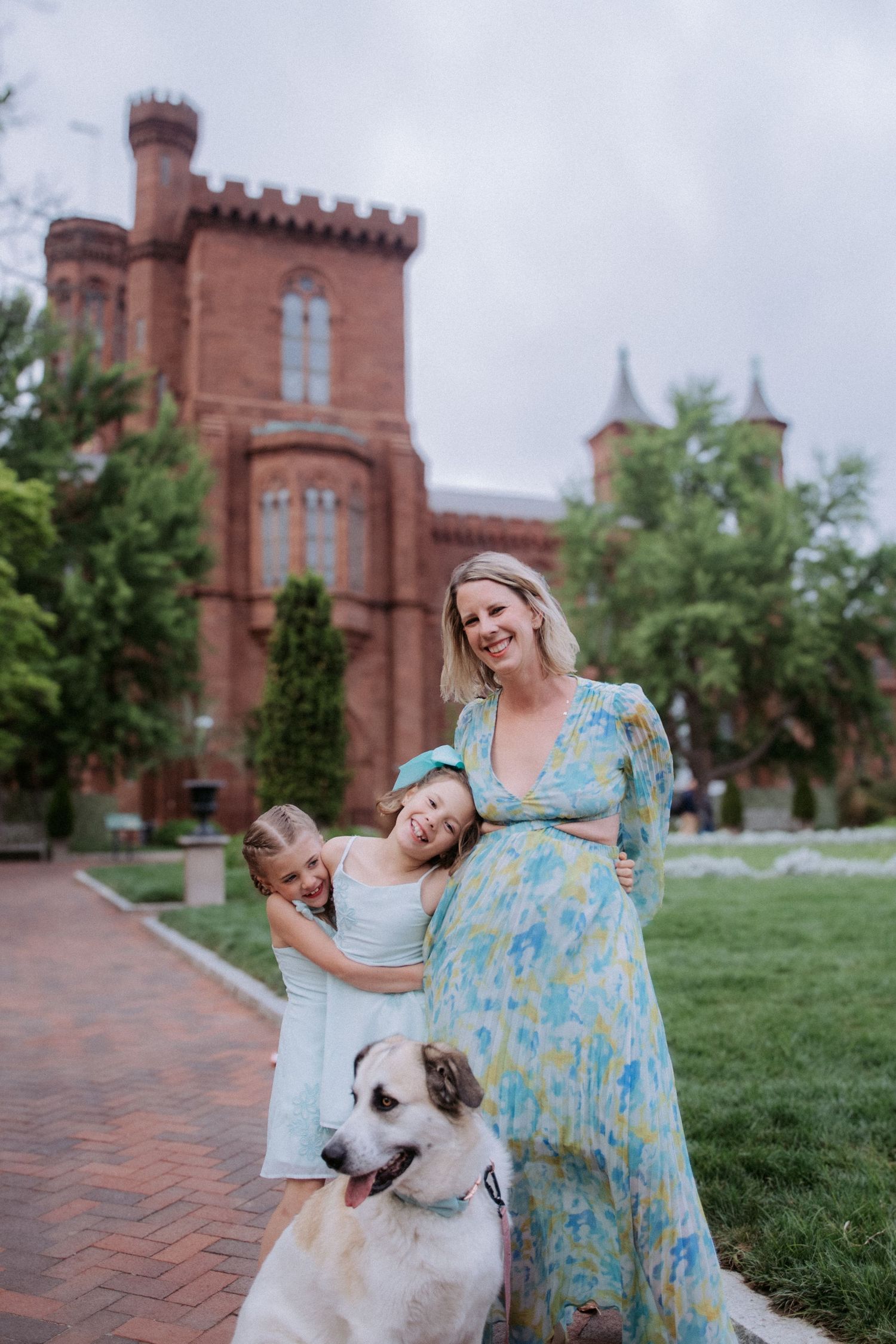 Pregnant woman in floral dress poses with two daughters and a dog in front of a red brick castle.