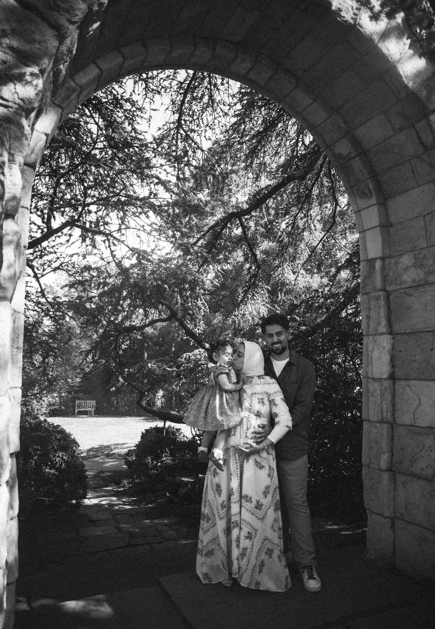 Black and white photo of a couple standing under a stone arch surrounded by trees in a garden setting.