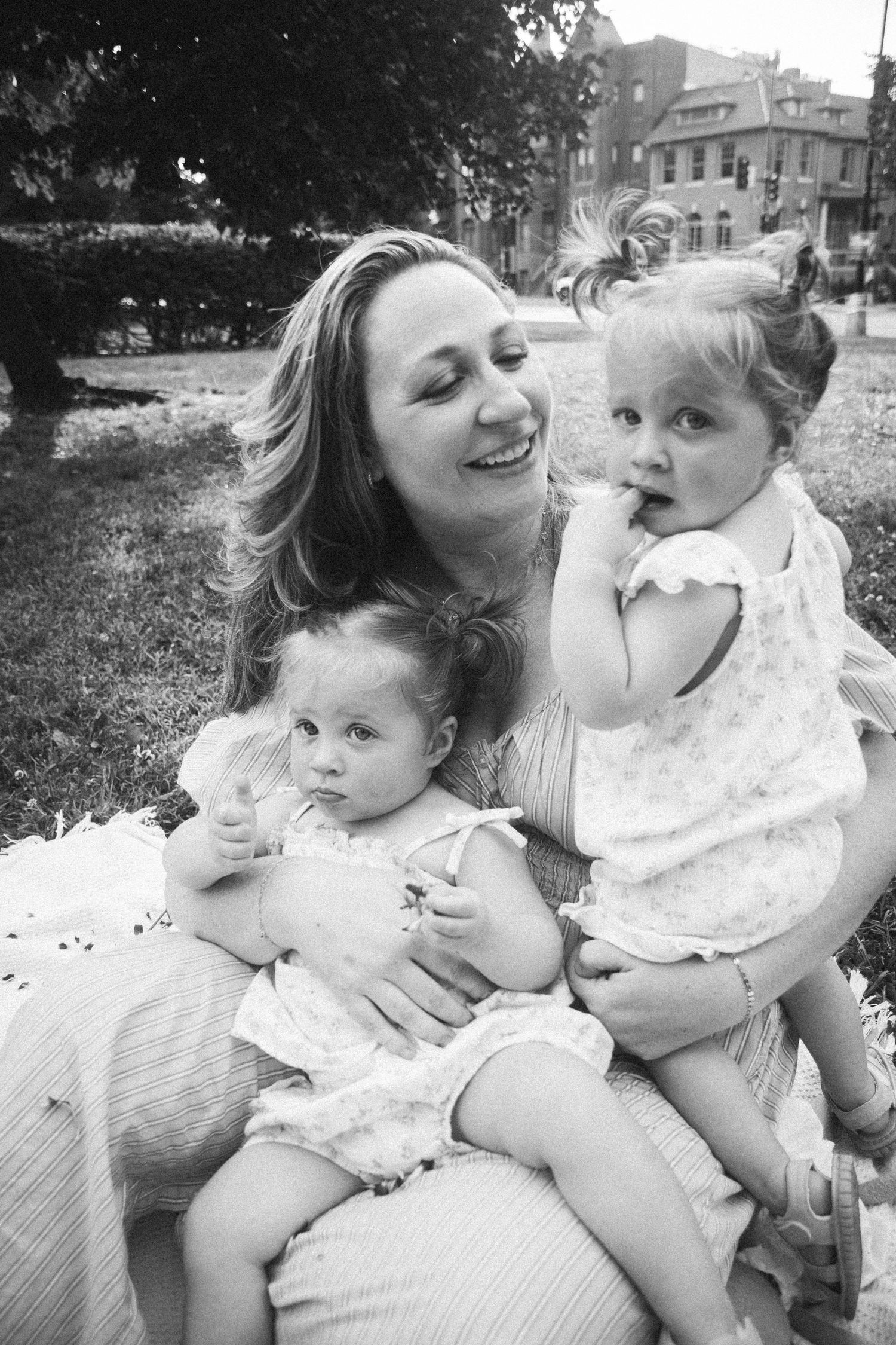 Black and white photo of a smiling mother sitting outdoors holding two young toddler girls on her lap in a park.