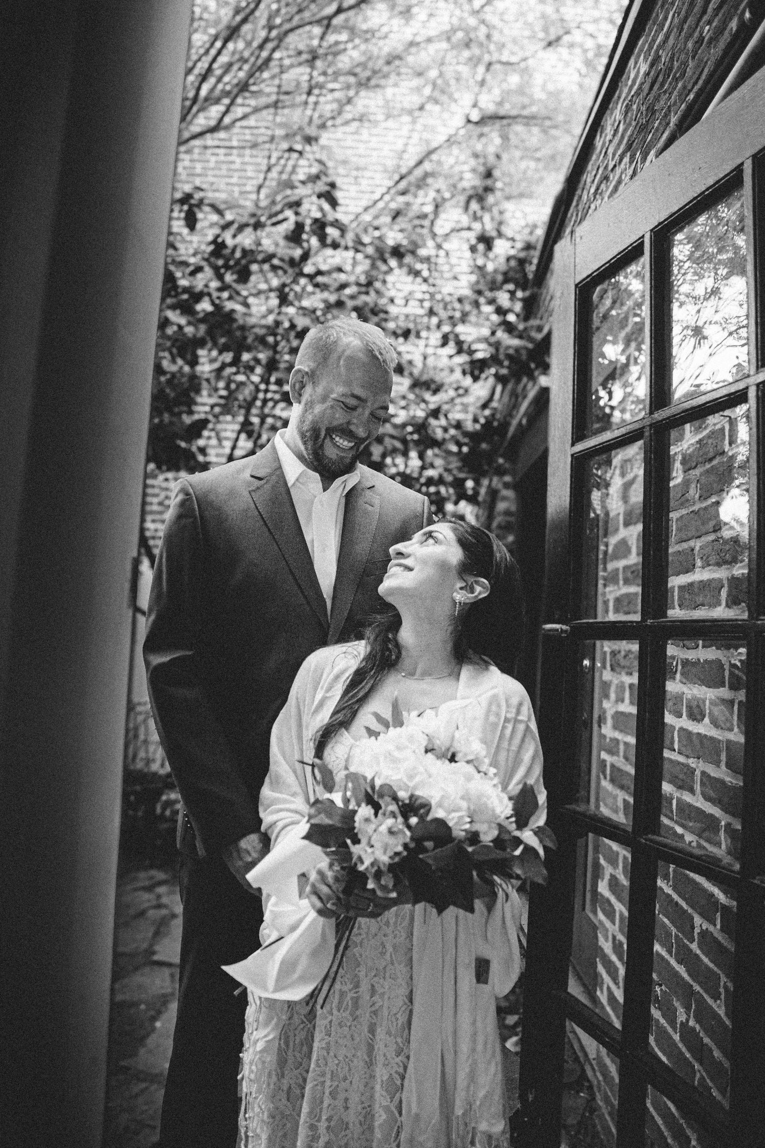 Black and white wedding photo of smiling bride holding bouquet and groom sharing a joyful moment by brick wall.