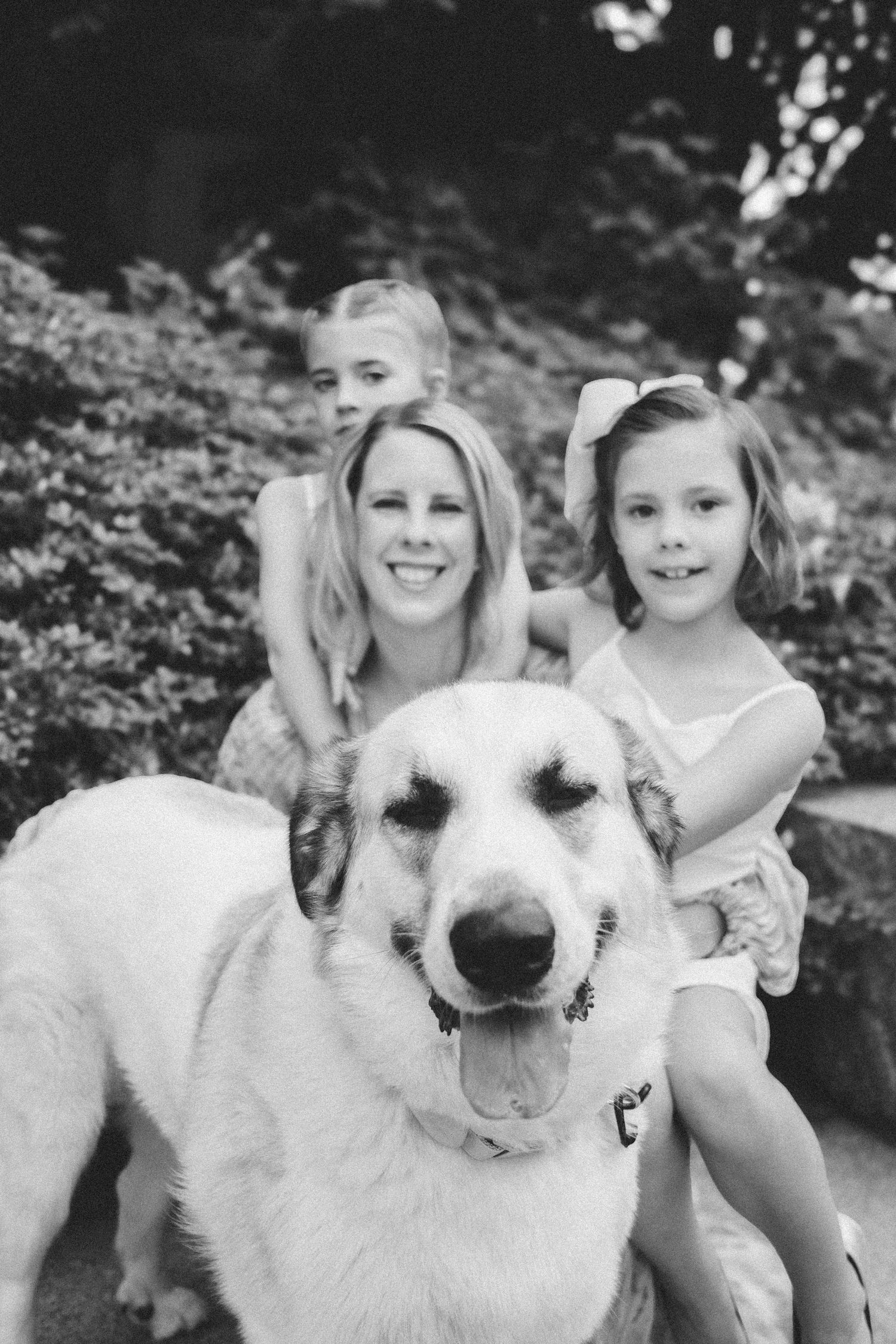 Black and white photo of a smiling woman with two young girls and a large white dog outdoors.