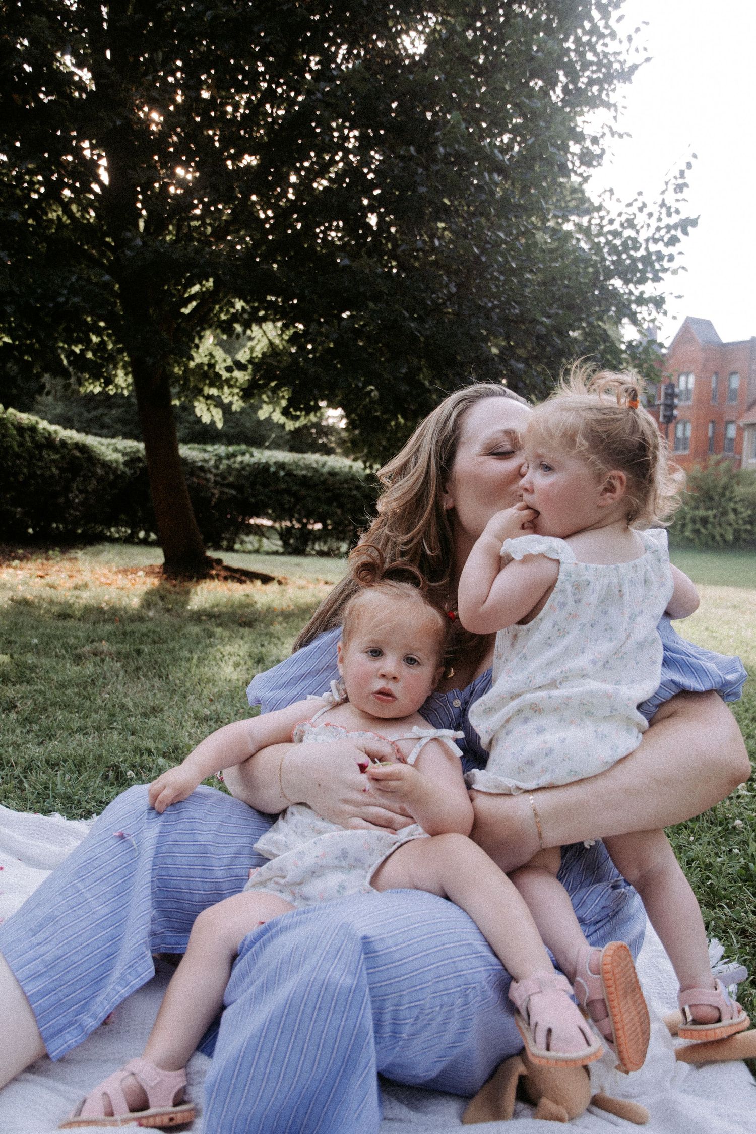 A mother sits on a blanket in a park, kissing her toddler while holding a baby on her lap.