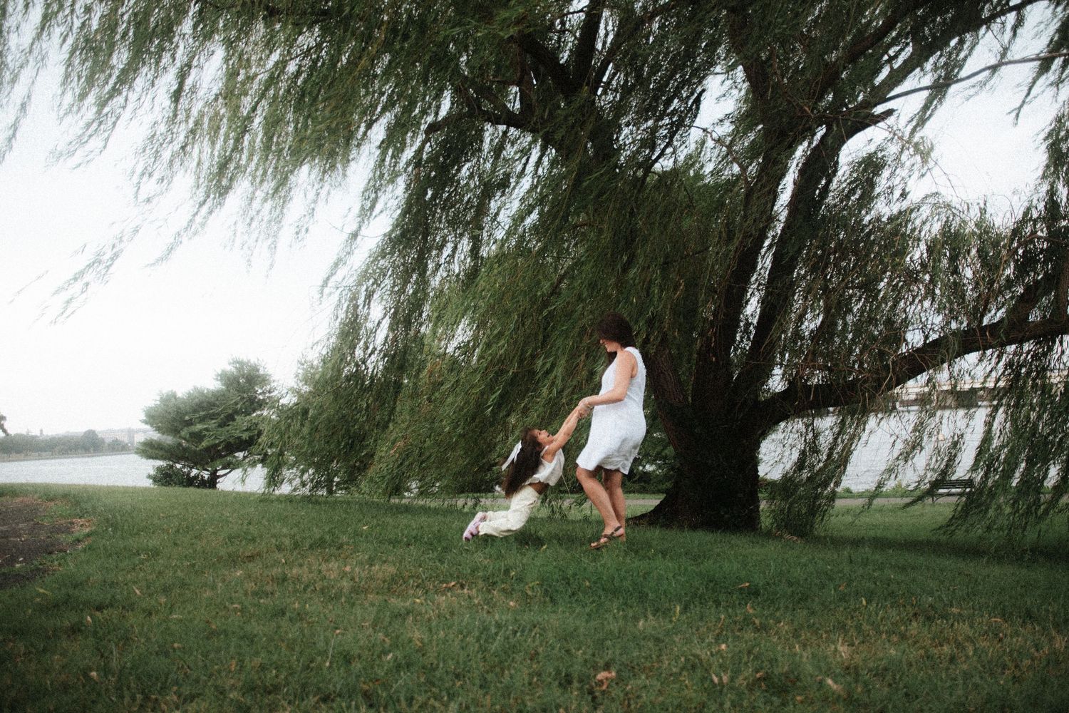 A woman in a white dress plays with a child under a weeping willow tree by a lake on a green lawn.
