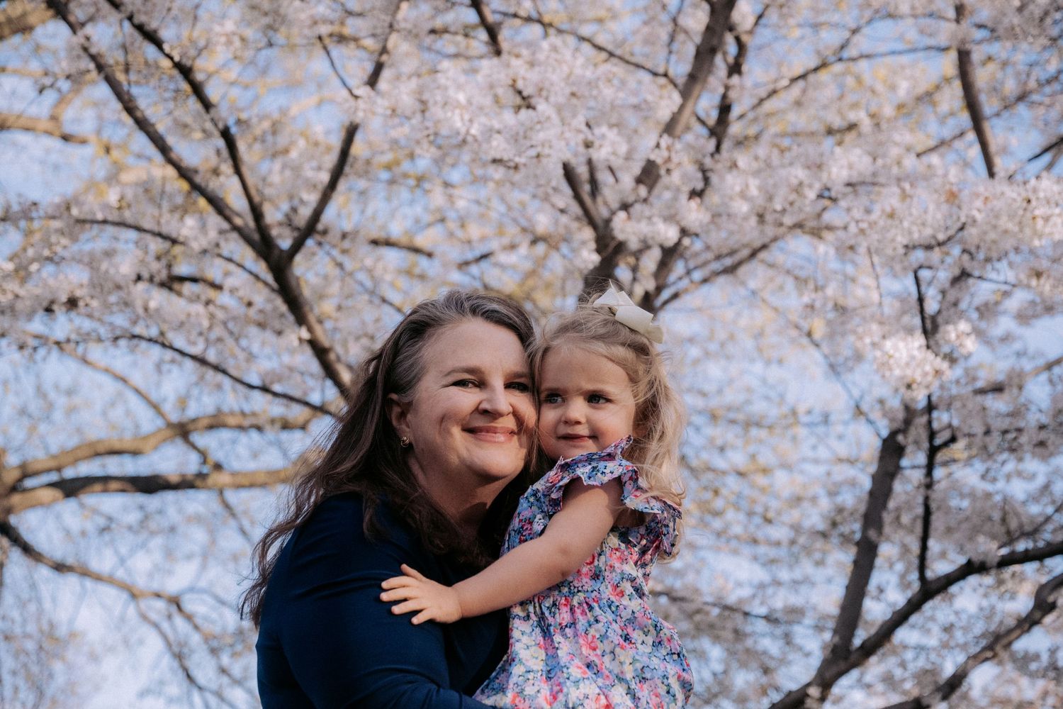 A smiling woman holds a young girl in a floral dress under blooming cherry blossom trees in spring.