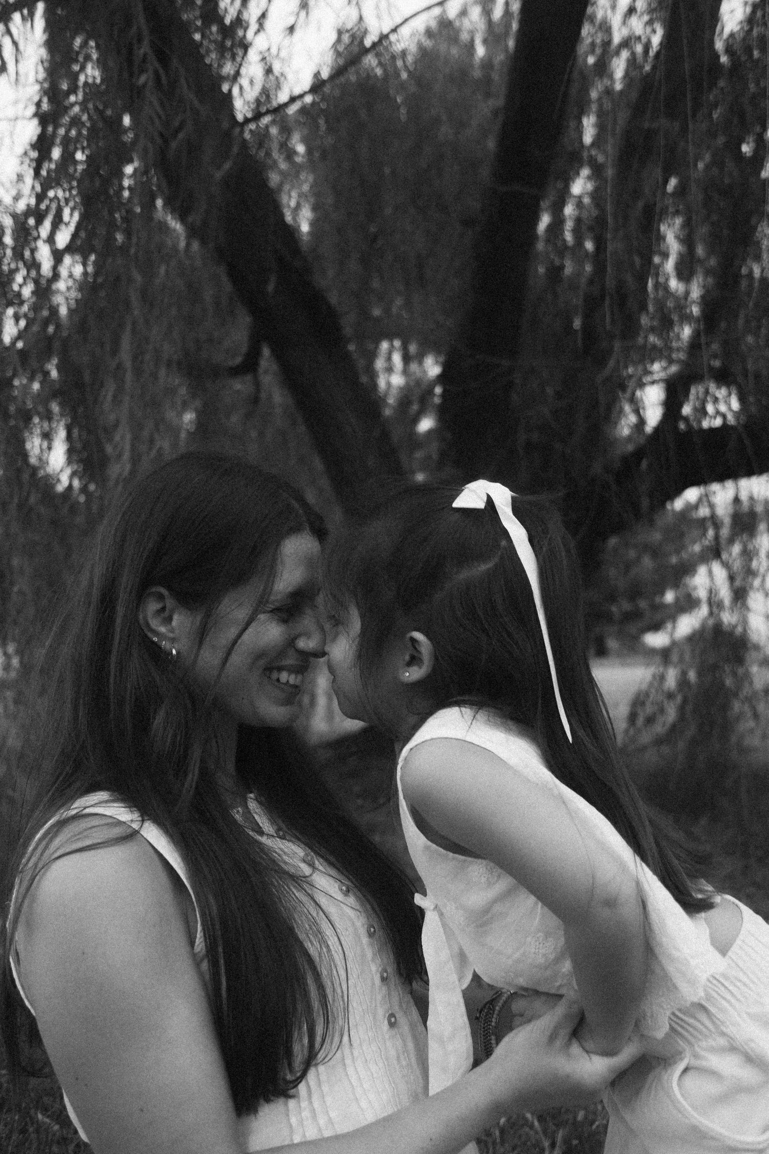 Black and white photo of a smiling mother holding her young daughter in a white dress outdoors near a tree.