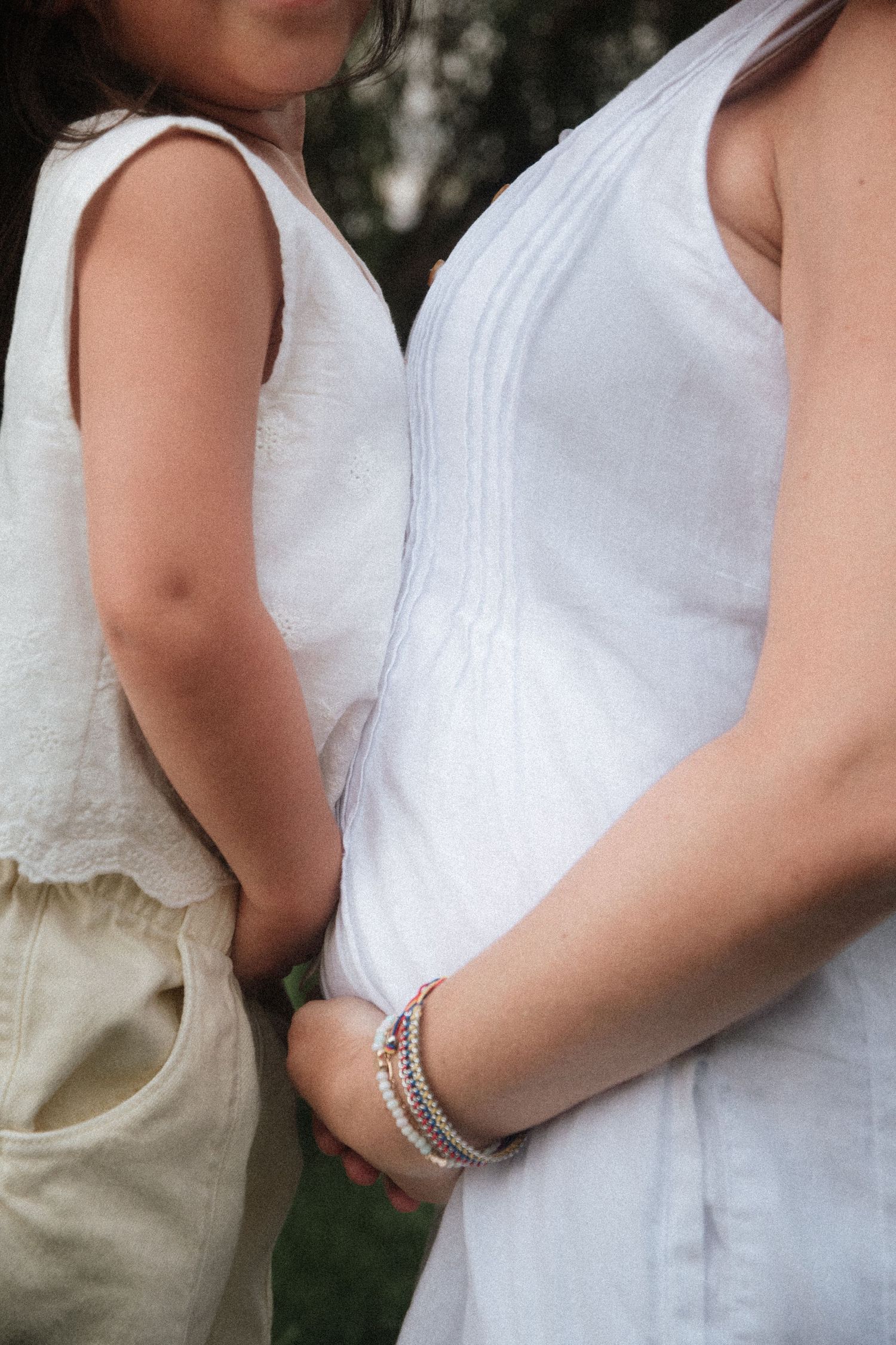 Pregnant woman in white top holding hands with young child in white dress outdoors.