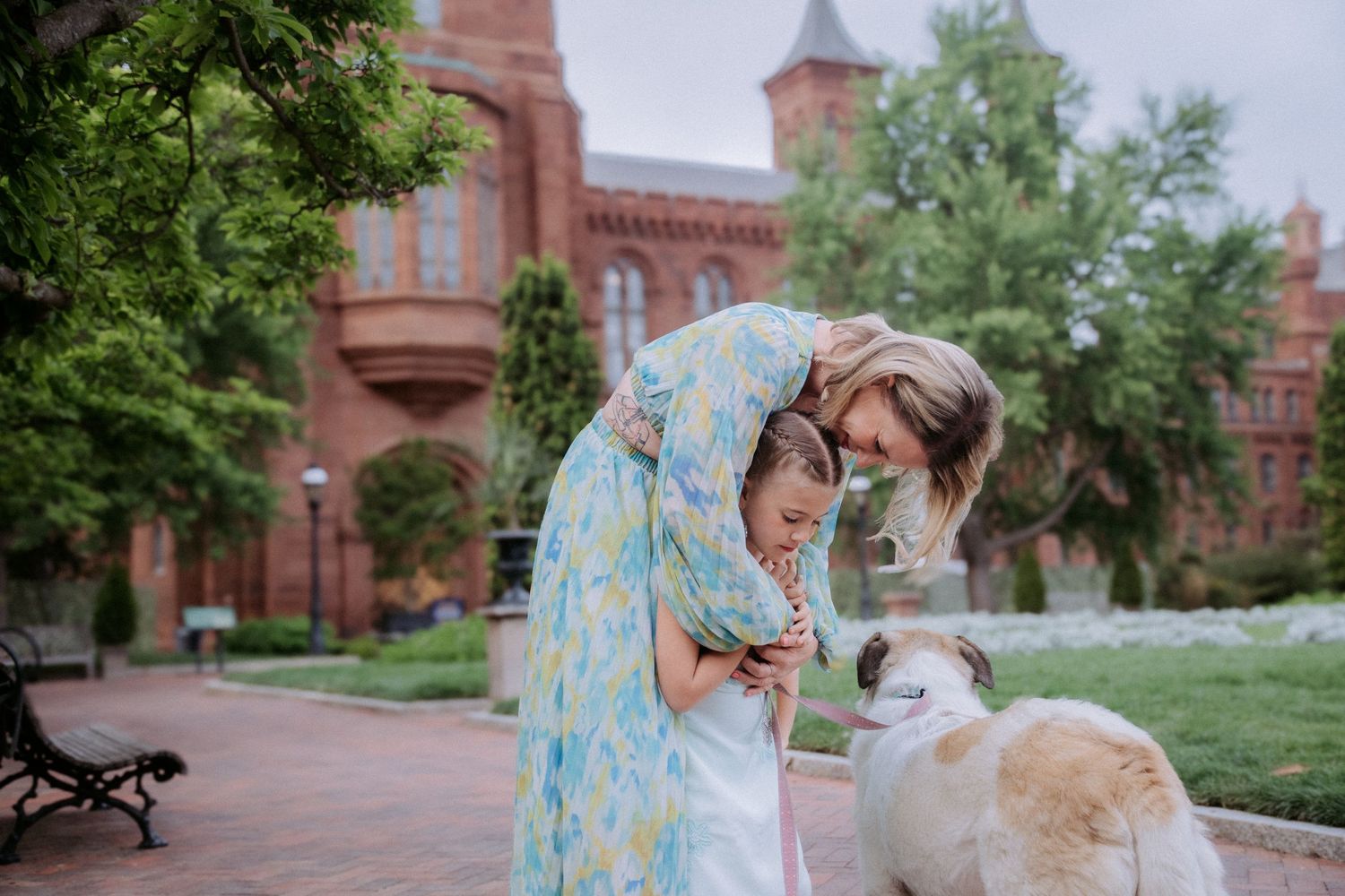 A mother and daughter bond over a dog outdoors near a red brick building in a lush green garden.
