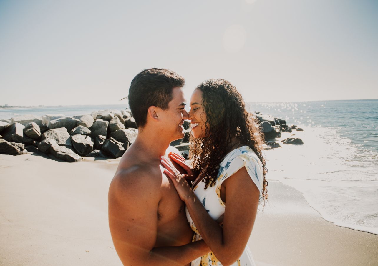 Romantic couple embracing on a sunny beach with ocean waves and rocks in background.