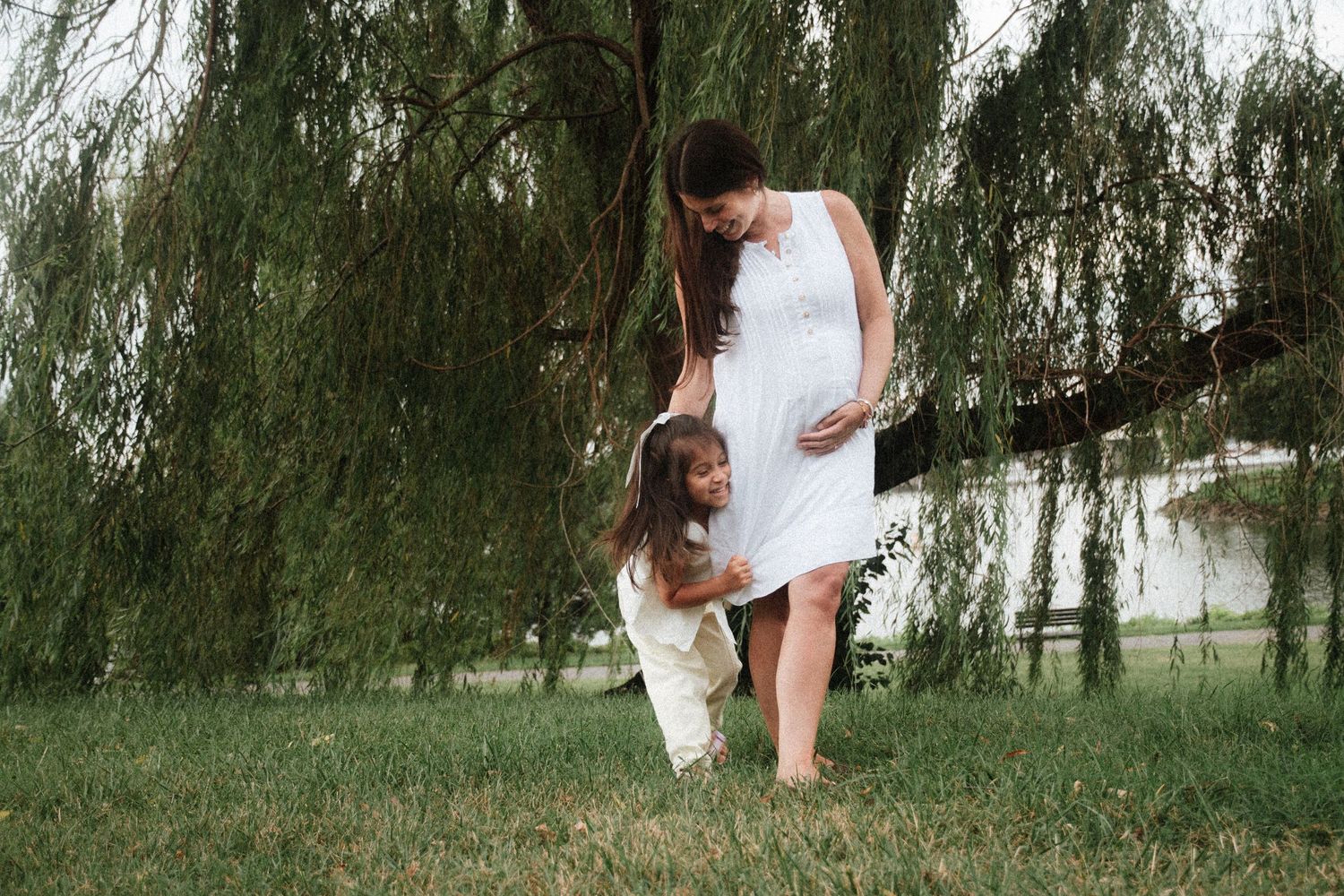 Pregnant woman in white dress with young daughter touching her baby bump near a weeping willow tree outdoors.