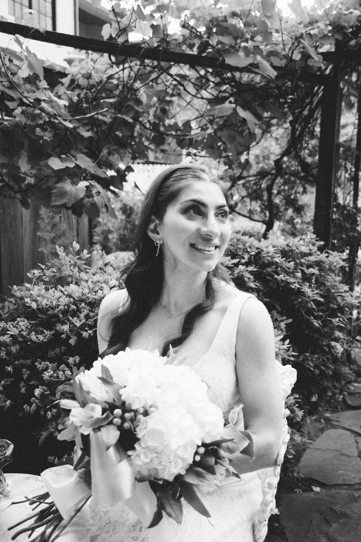 Black and white photo of a smiling bride holding a white floral bouquet, standing outdoors under a leafy garden arbor.