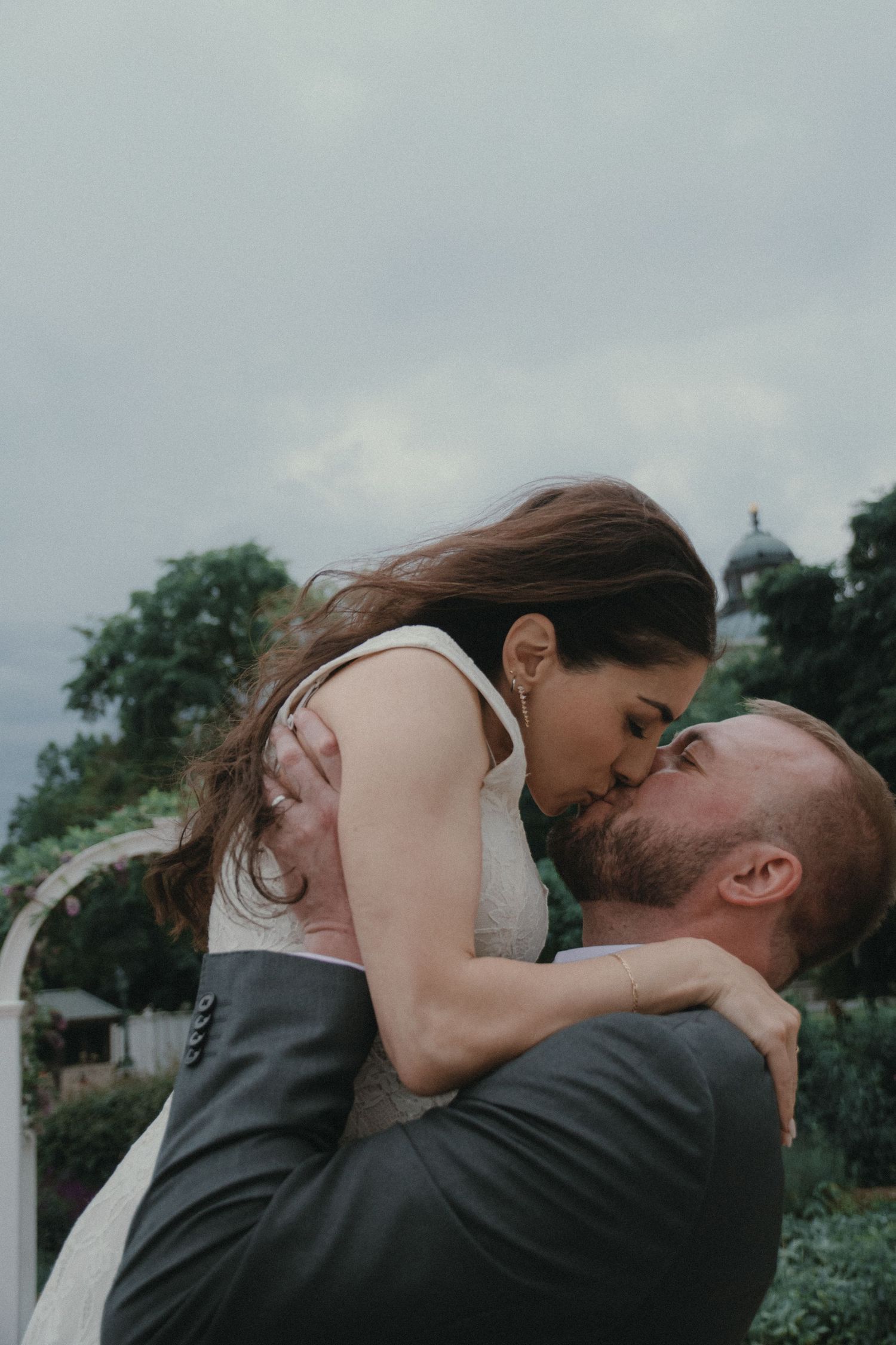 Groom lifts bride for a romantic kiss outdoors at a garden wedding venue under a cloudy sky.