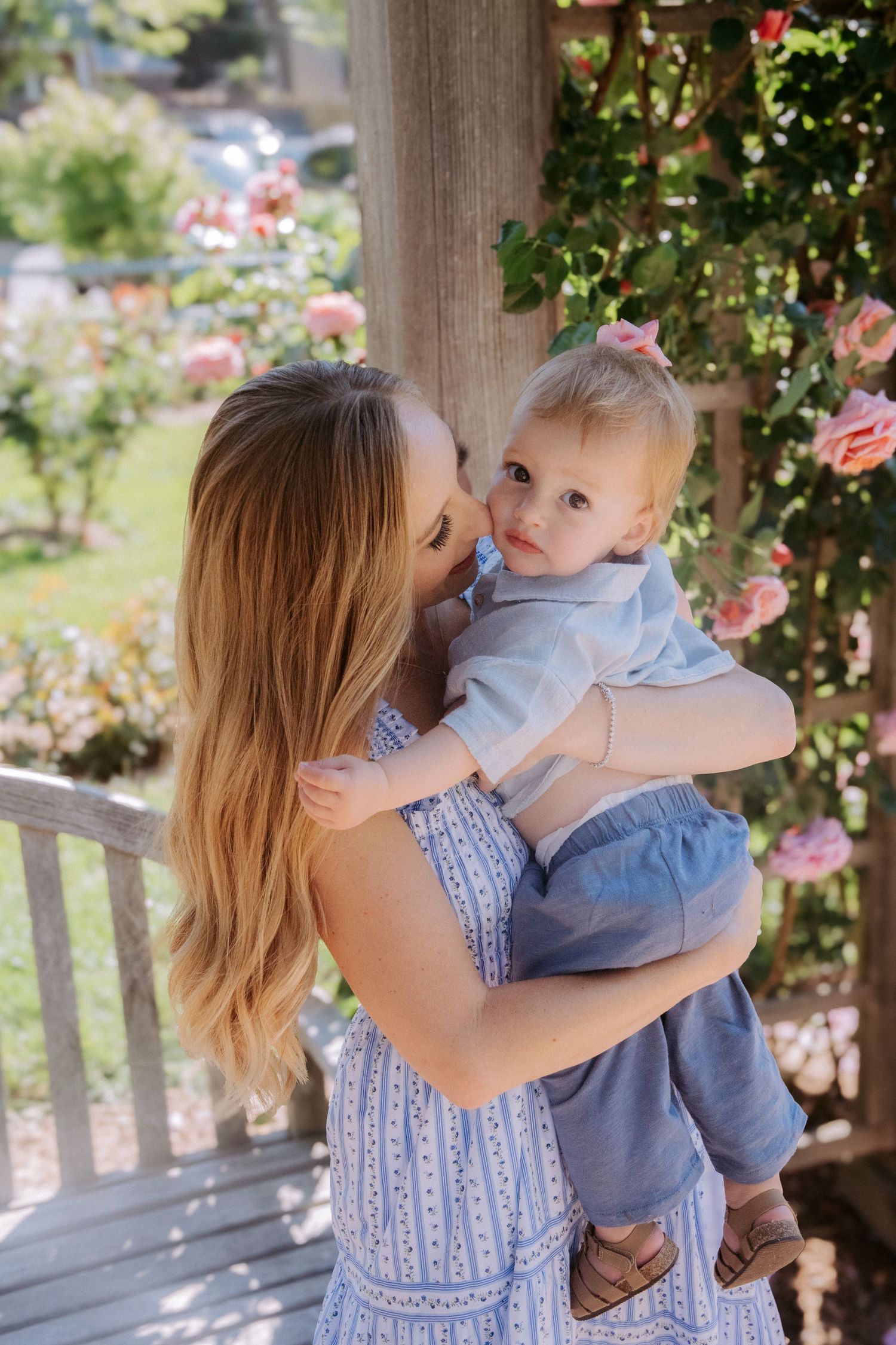 A mother in a blue dress holds her baby girl near a rose-covered trellis outdoors.