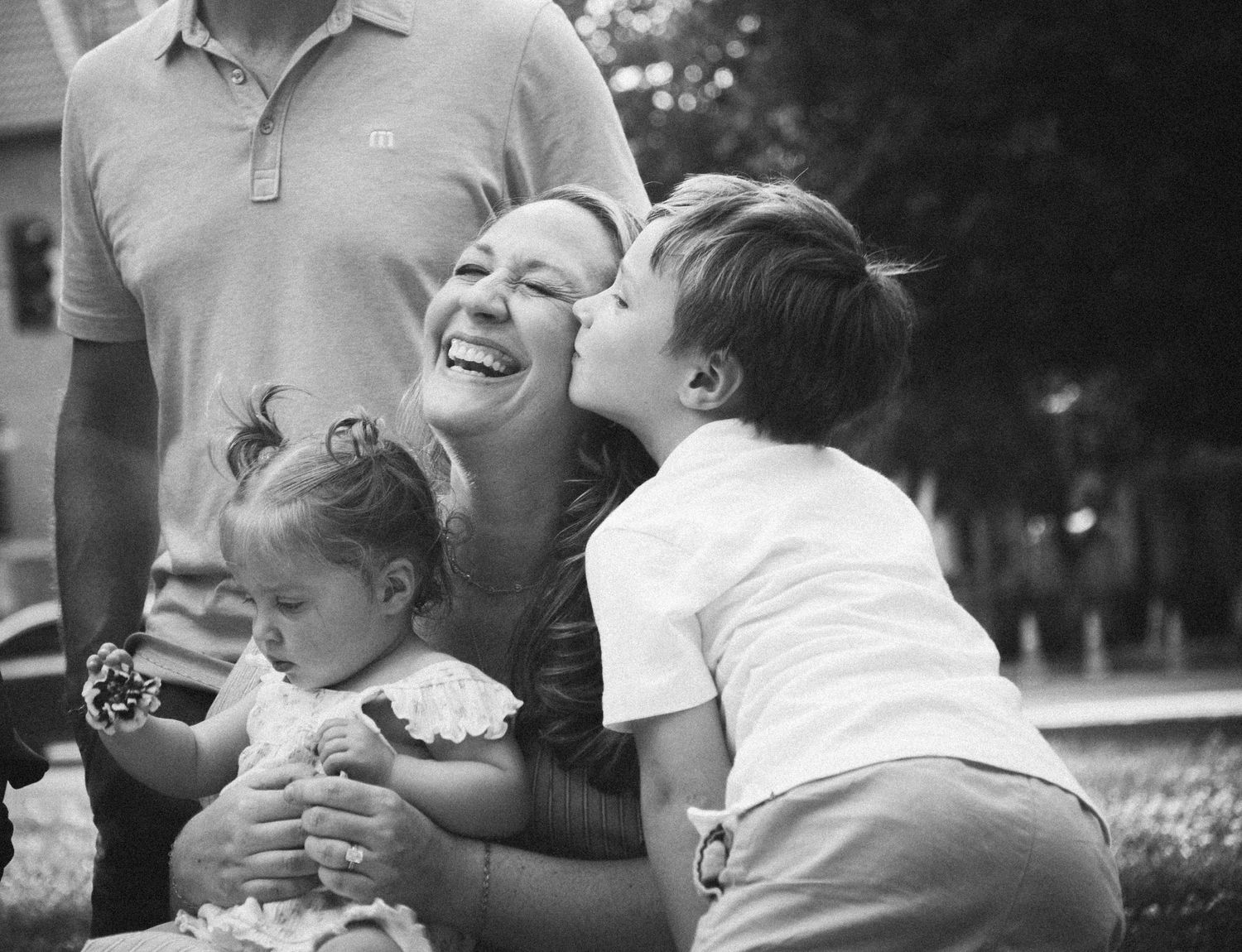 Black and white photo of a laughing mother being kissed by her son while holding a baby girl outdoors.