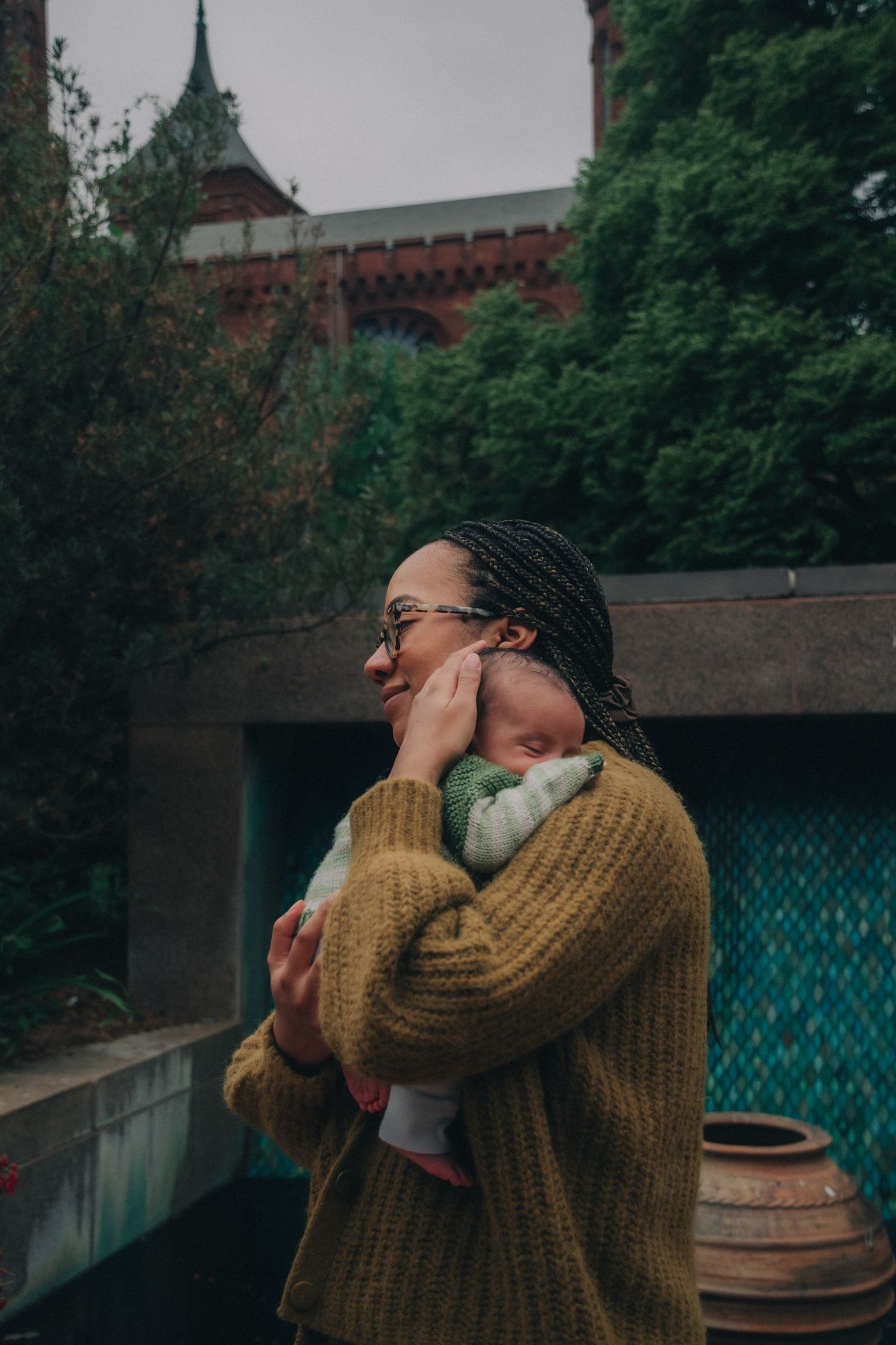 A woman with braids and glasses holds a newborn baby outdoors near a brick building surrounded by lush greenery.