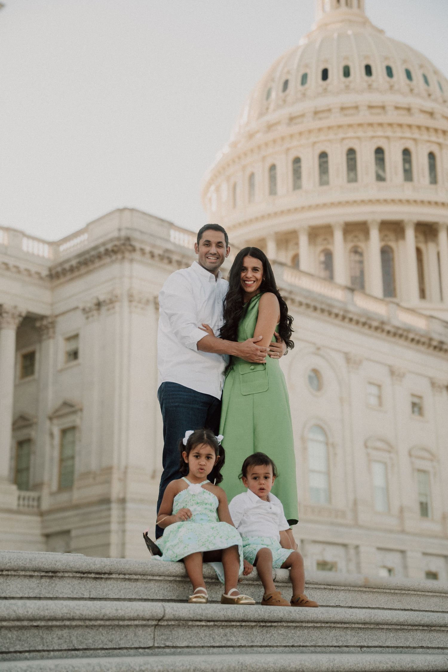 Family of four posing on steps of US Capitol building, parents embracing behind two young children in coordinating outfits.