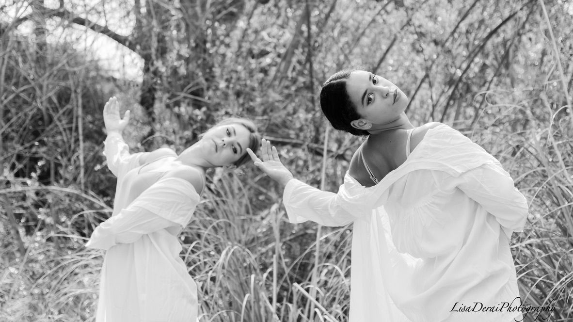 Dancer in white dress poses gracefully against a natural outdoor backdrop with trees and foliage.