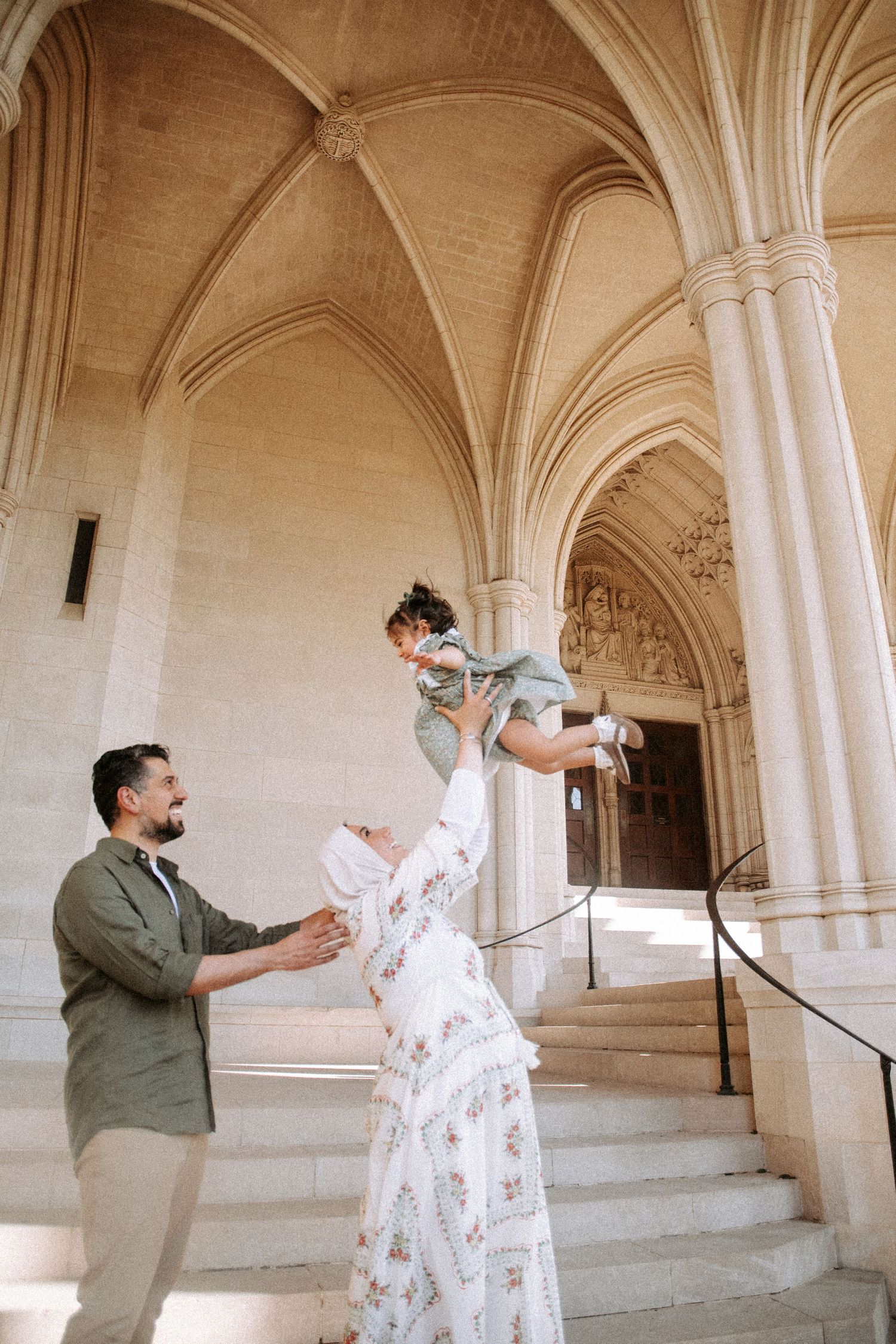 Family portrait with mother lifting baby in Gothic cathedral archway, father watching joyfully.