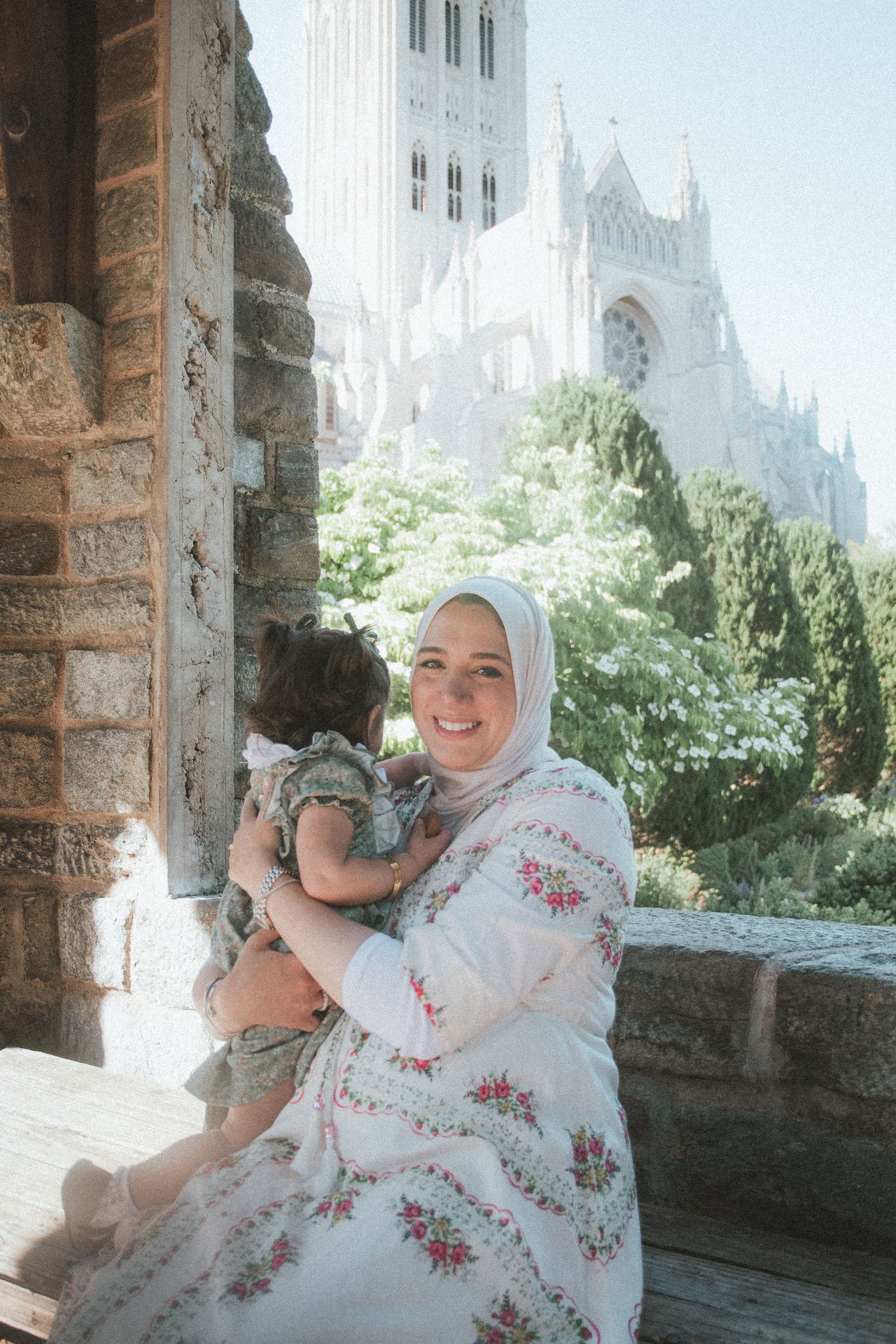 Smiling Muslim mother in hijab holding baby girl outside Gothic cathedral on sunny day.