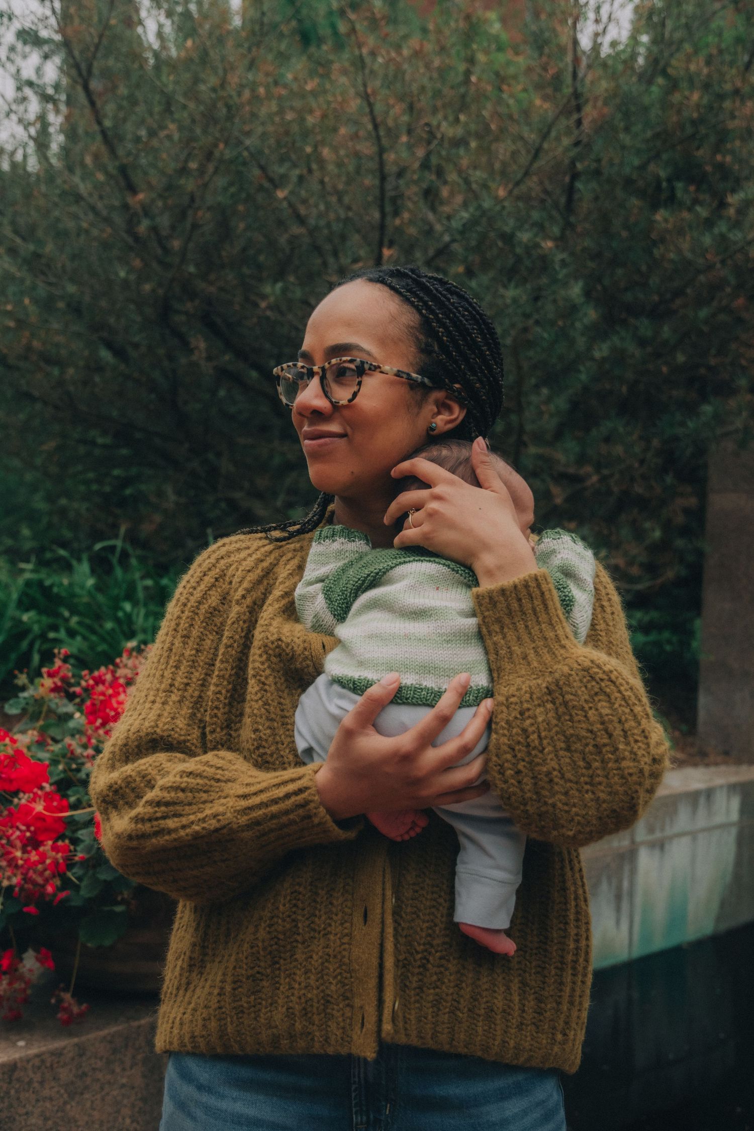 A smiling woman with braids and glasses holds a baby outdoors, wearing a mustard knit cardigan near garden greenery.