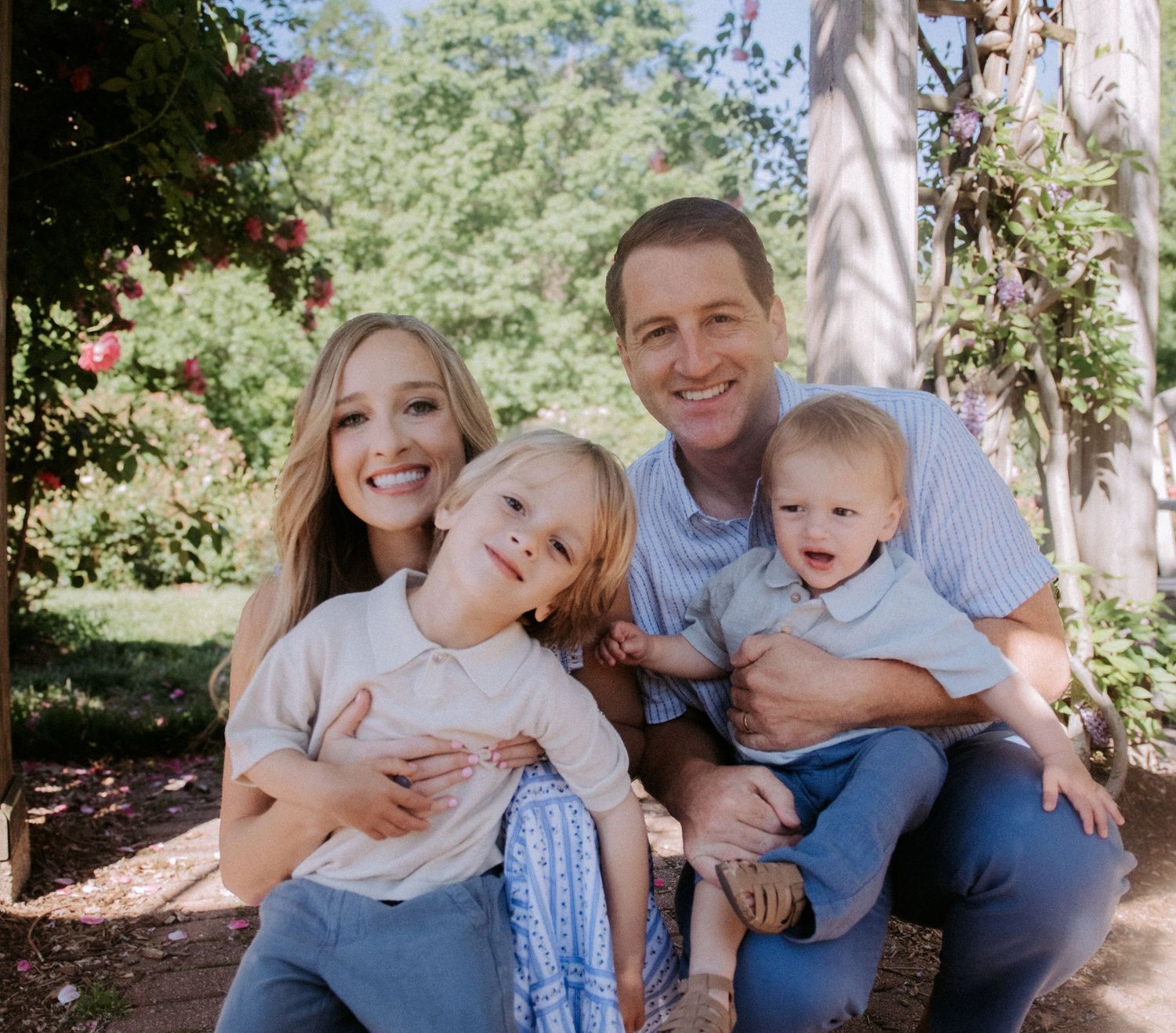 Happy family of four posing outdoors in a lush garden, parents smiling with two young children.