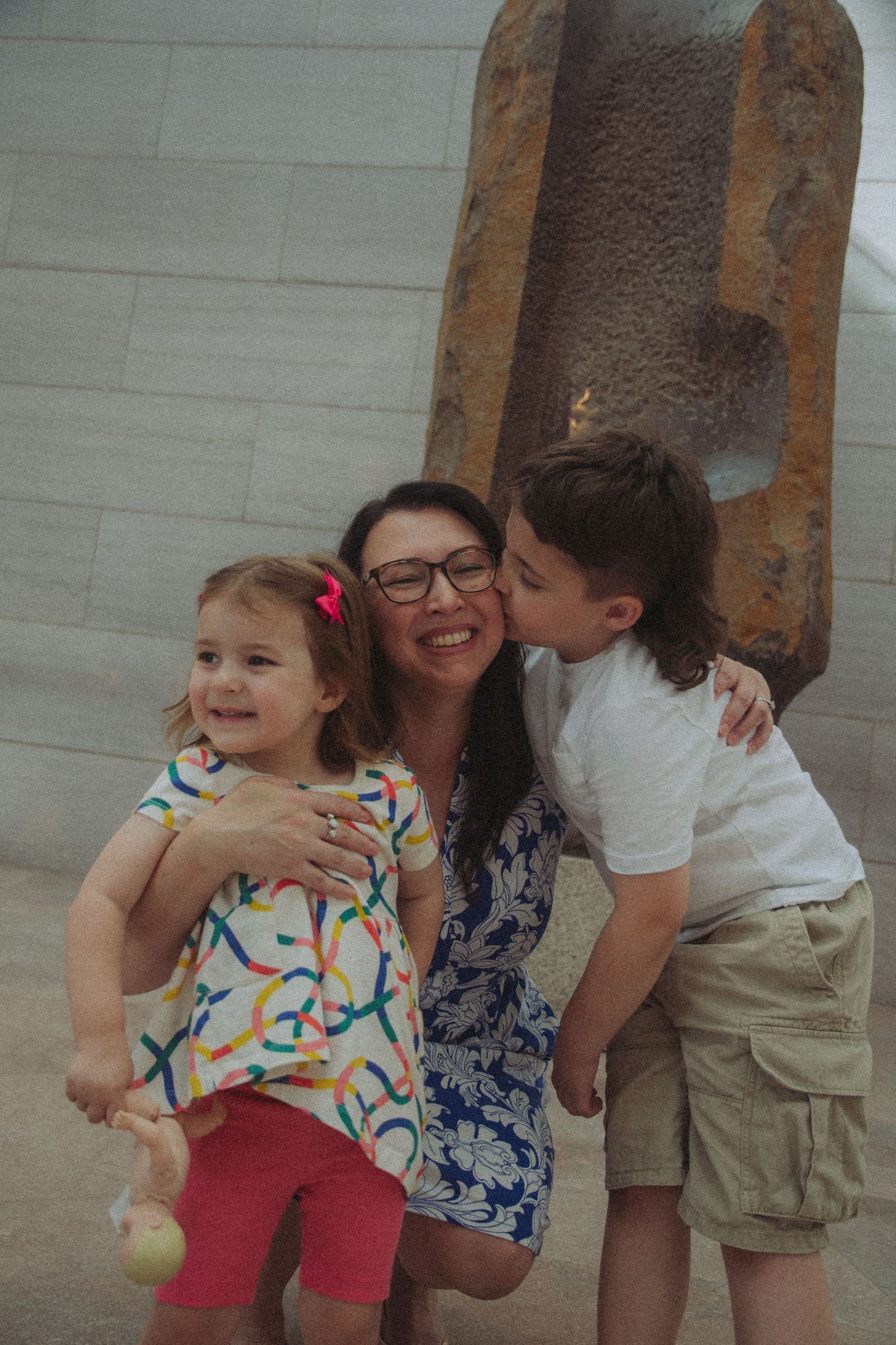 A smiling woman with glasses holds a toddler girl while a boy kisses her cheek outdoors near a stone sculpture.